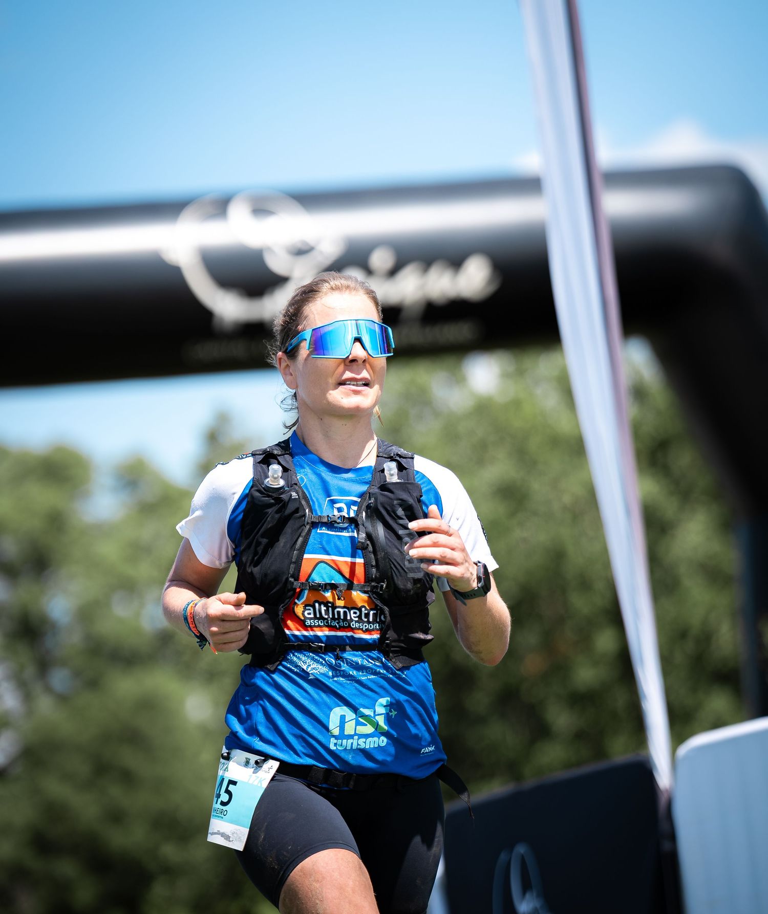 Runner wearing blue sunglasses and race vest smiles during outdoor trail running event.