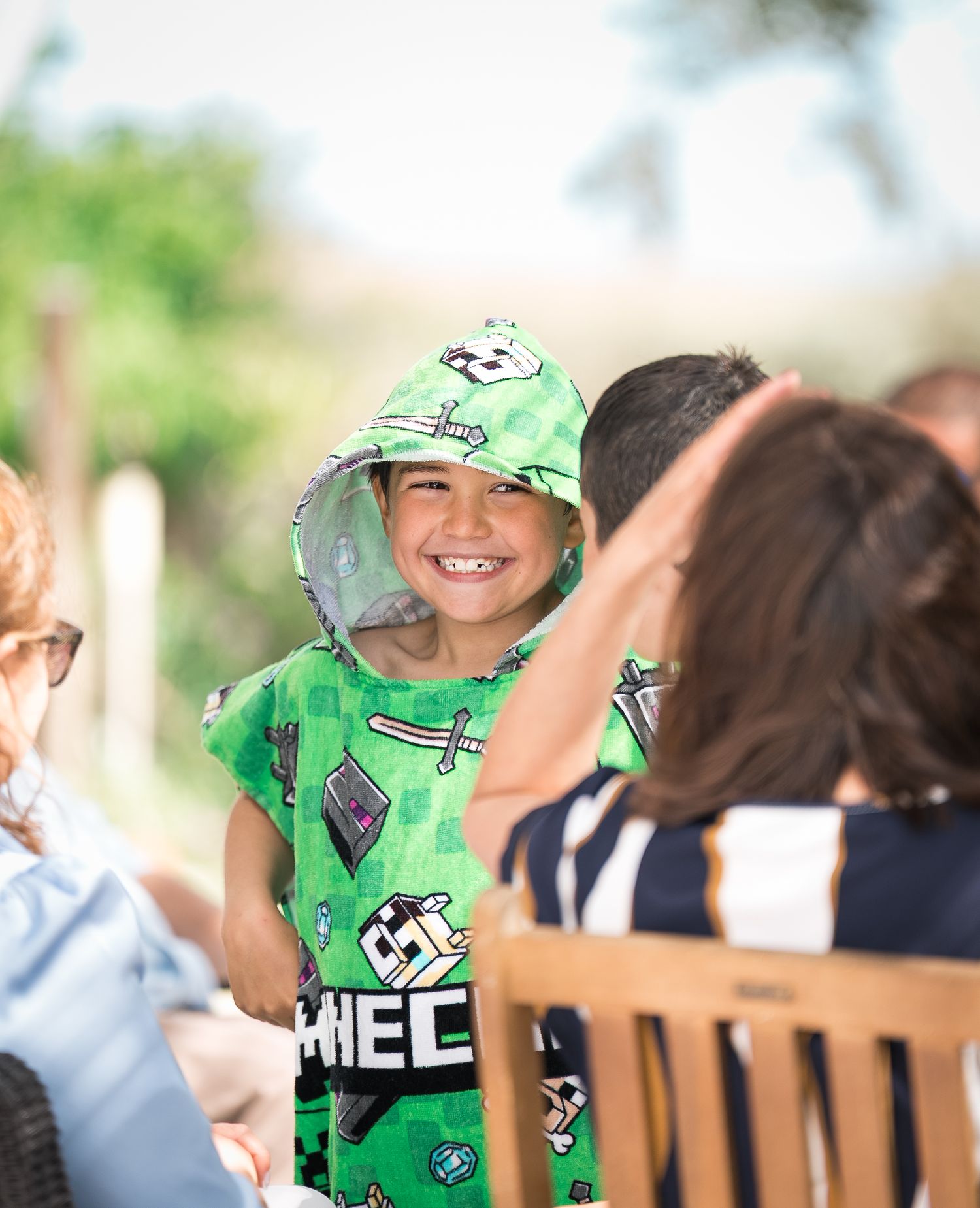Child wearing green patterned outfit and hat smiling at outdoor gathering.