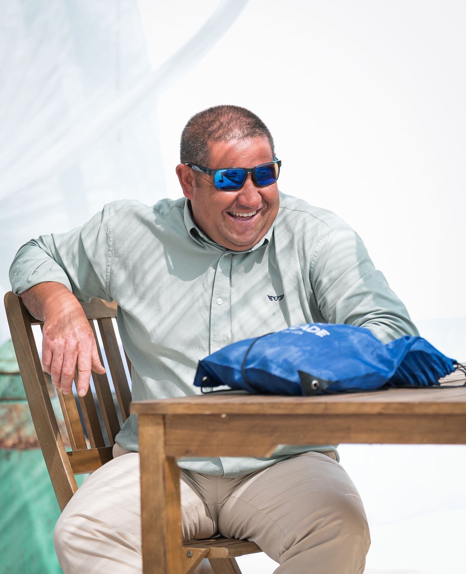 Person in sunglasses and light shirt relaxing at outdoor table with blue bag.