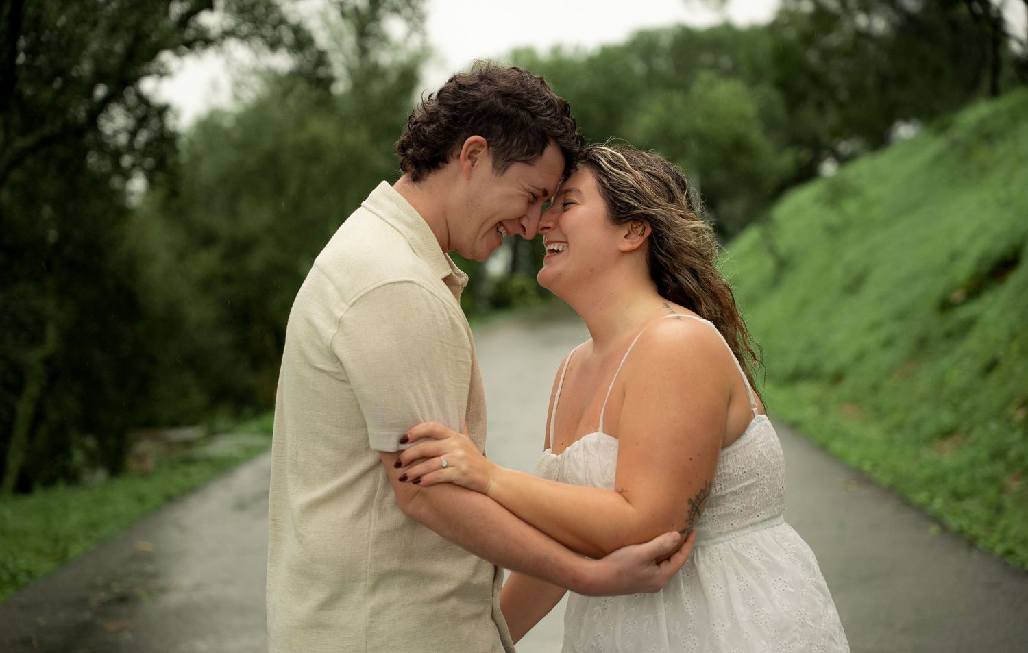A couple embraces and touches foreheads romantically on a tree-lined path, the woman in a white dress and man in a beige shirt.