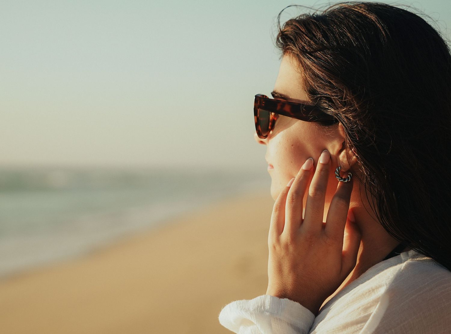 Side profile of woman in sunglasses touching face near ocean.