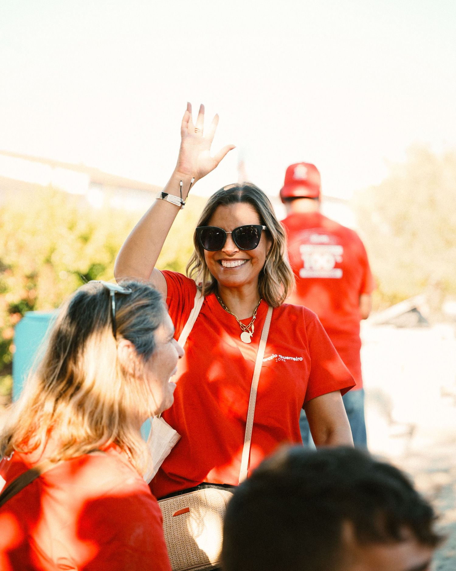 A cheerful outdoor gathering with people in red shirts and sunglasses waving to the camera.