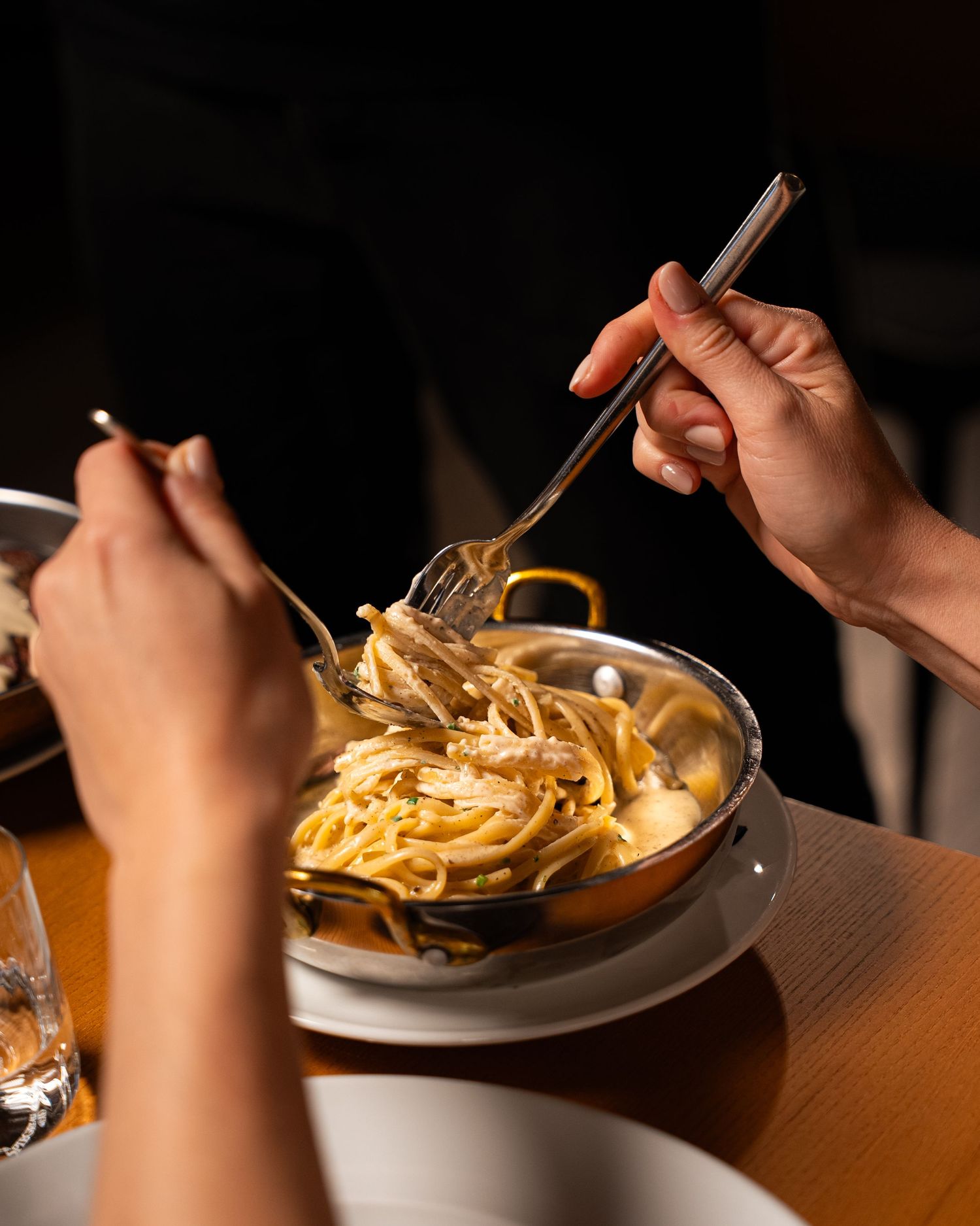 Two hands twirling creamy fettuccine pasta in a golden pan at a dimly lit restaurant table.