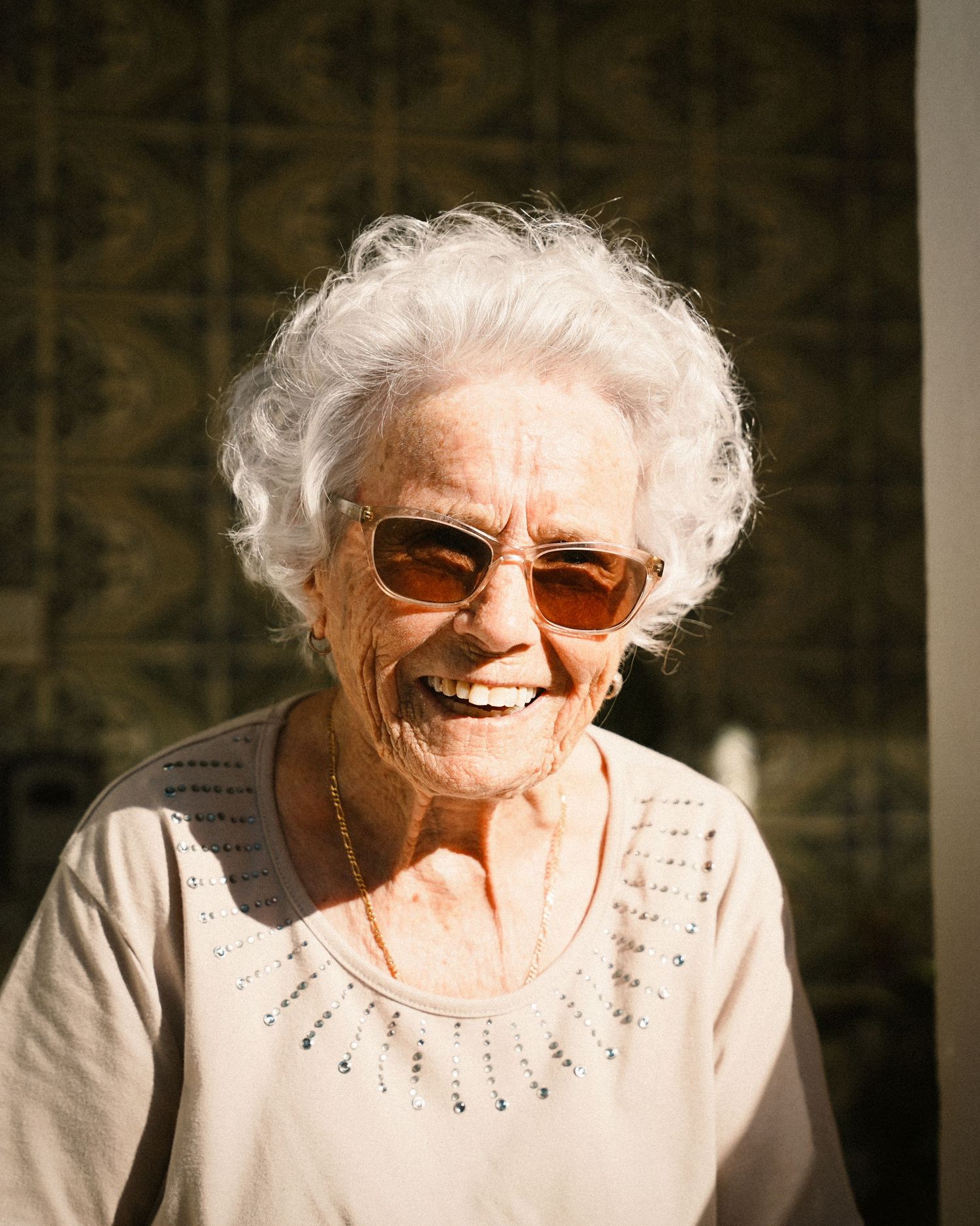 An elderly person wearing sunglasses smiles warmly in front of a patterned wall.