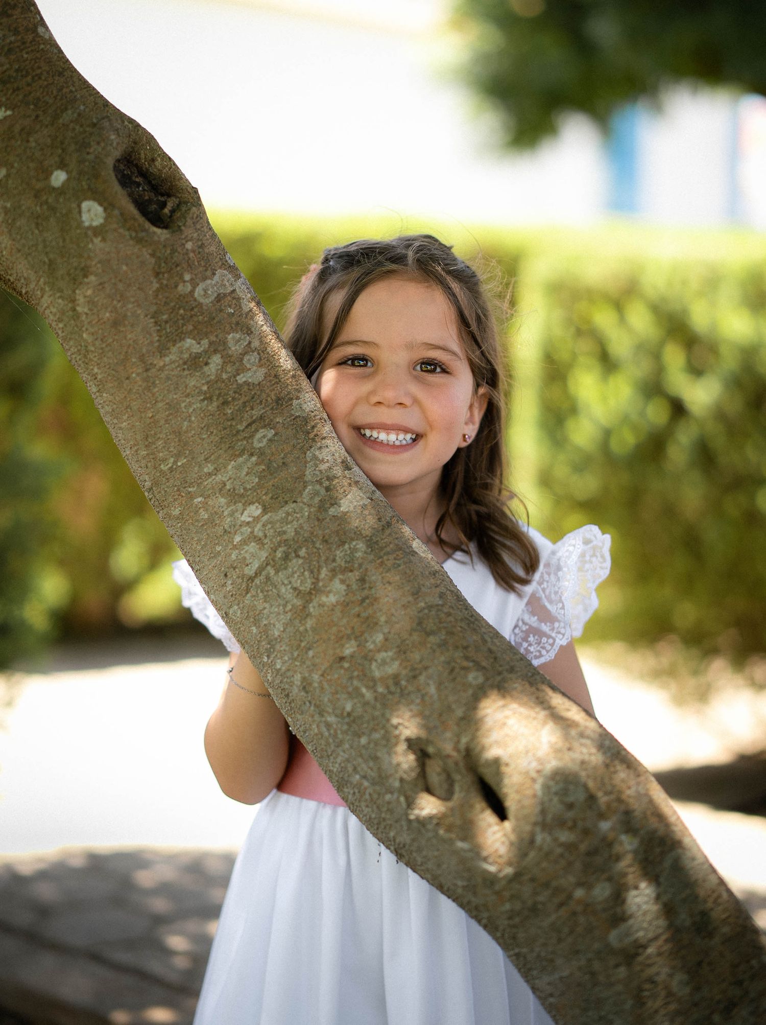 Young girl in white dress smiling while hugging a tree trunk outdoors in a sunny garden setting.