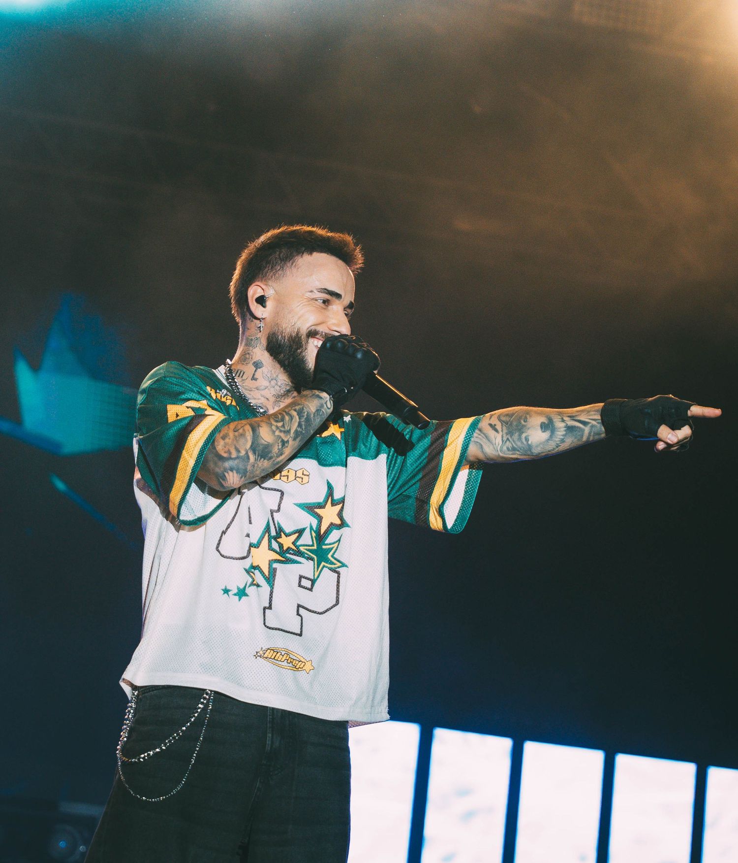 Performer in white jersey with tattoos points while singing on concert stage.