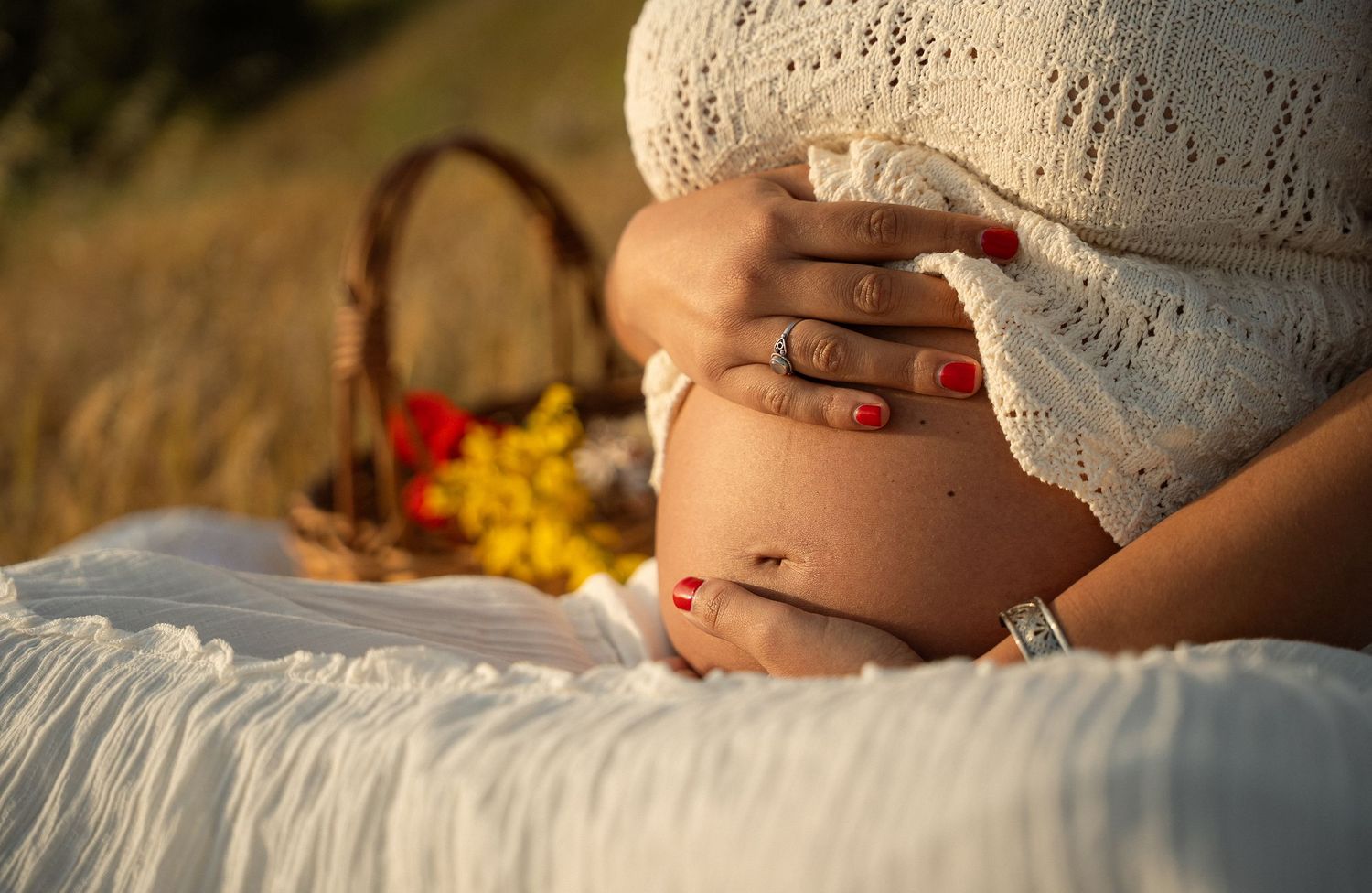 Pregnant woman in white lace dress cradling baby bump outdoors in golden field with picnic basket.