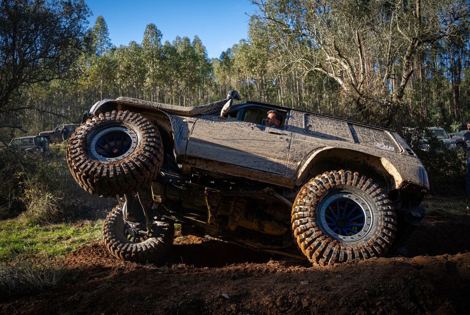 A heavily modified off-road vehicle with massive tires climbs over rough terrain in a wooded area.