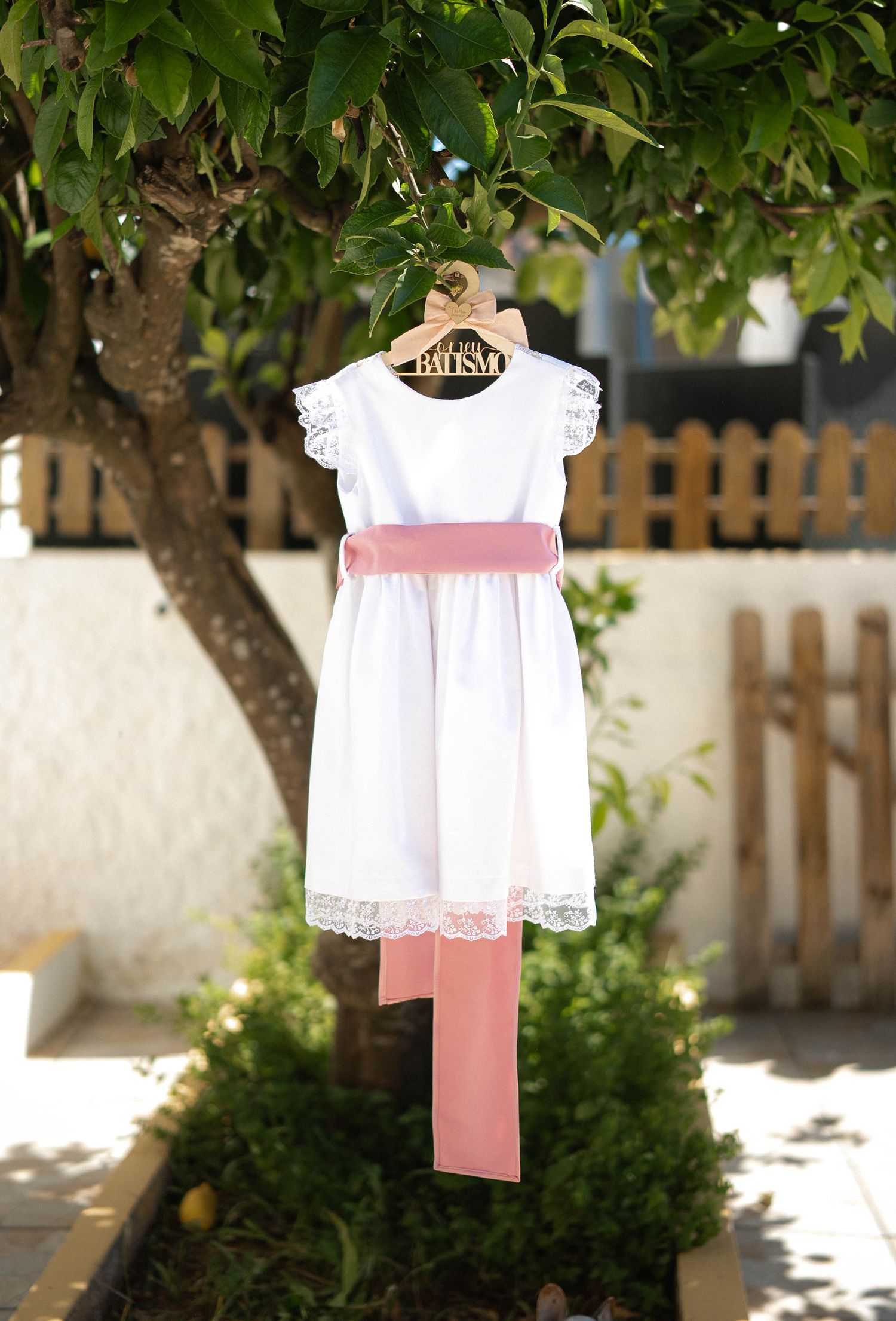 White lace baptism dress with pink satin sash hanging on personalized hanger from tree branch outdoors.
