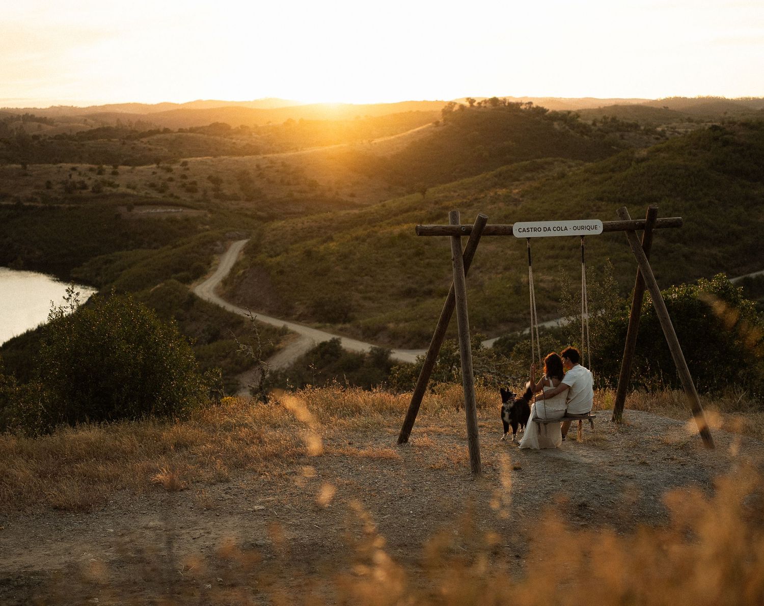 Couple sitting on a wooden swing at sunset overlooking a scenic valley with a winding path and river below.