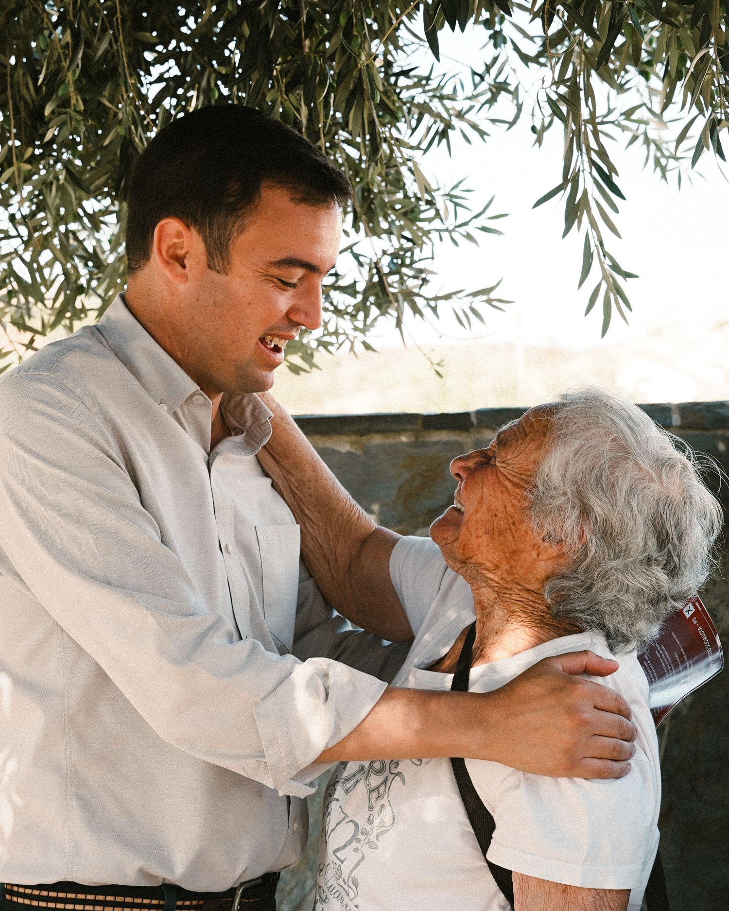 Two people sharing a warm embrace outdoors under olive branches.
