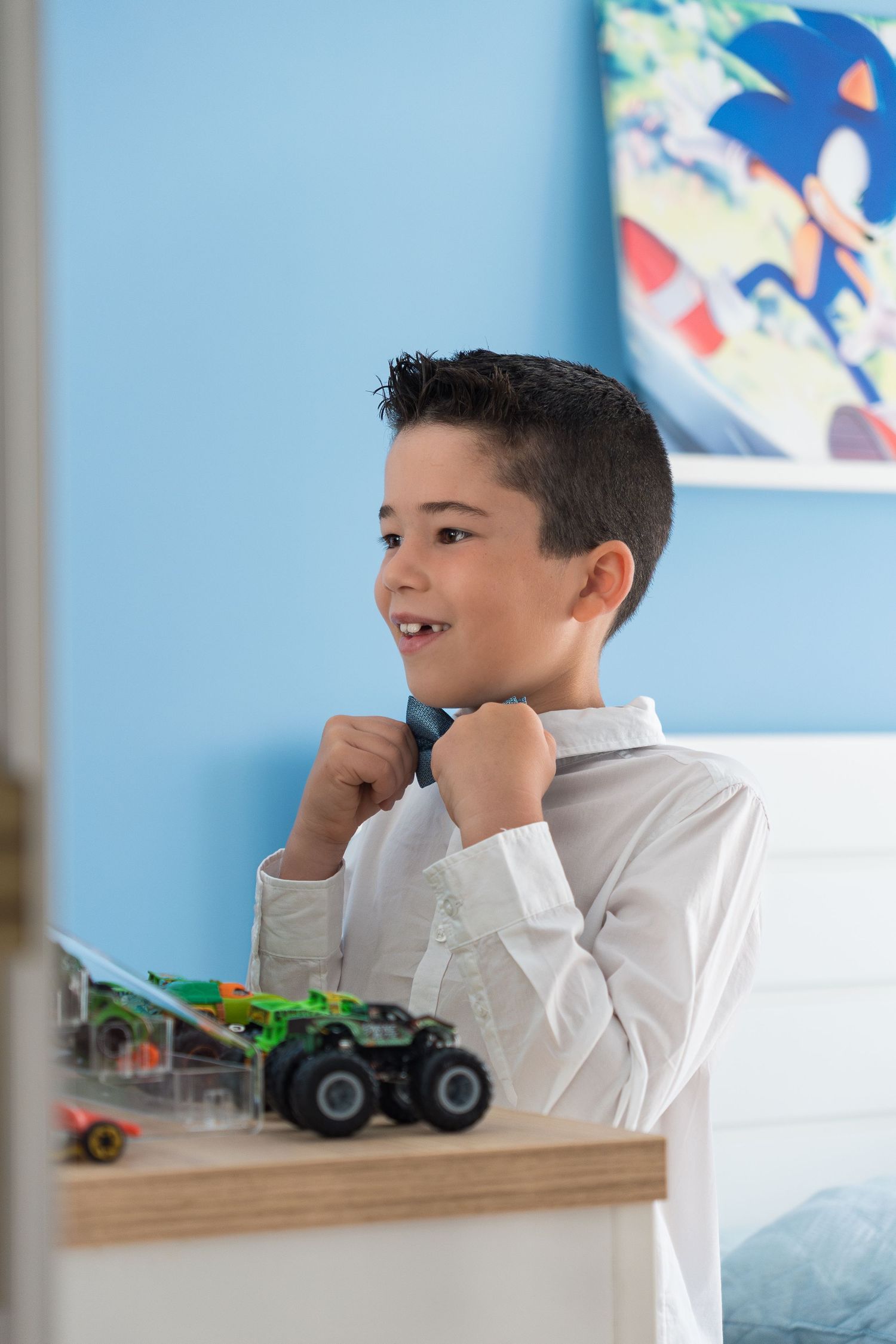 Young person adjusting bow tie while standing next to toy monster truck in bedroom.
