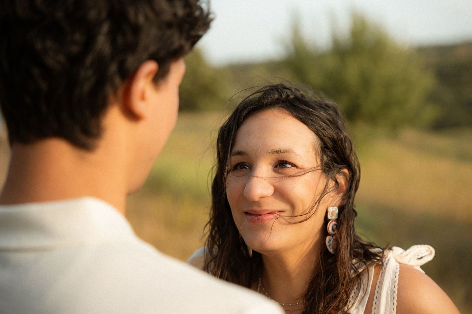 A young woman with dark hair smiles warmly at a man outdoors in golden hour sunlight.