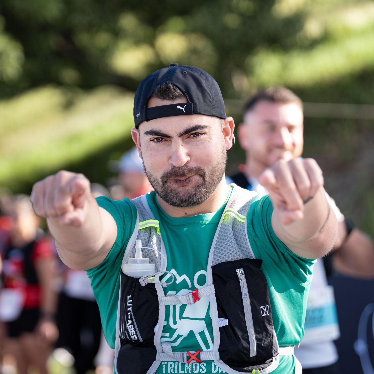 Trail runner in green shirt and hydration vest points at camera during race.