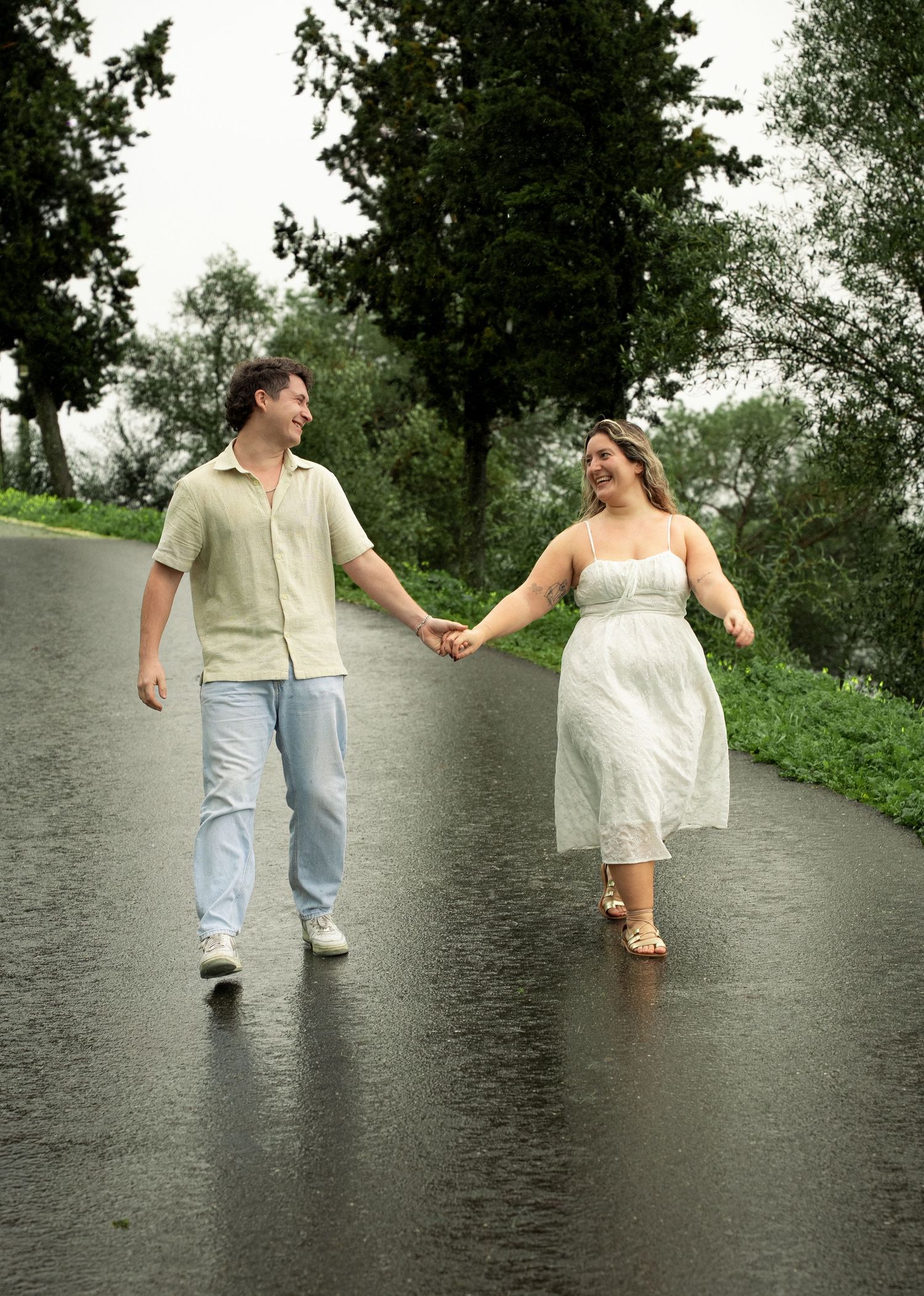 A couple holds hands and walks together on a rain-soaked road surrounded by lush green trees.
