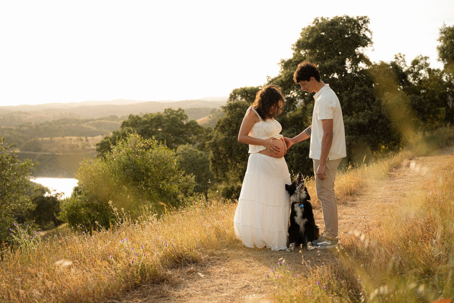Bride and groom holding hands with their dog on a scenic hilltop at golden hour with a lake view.