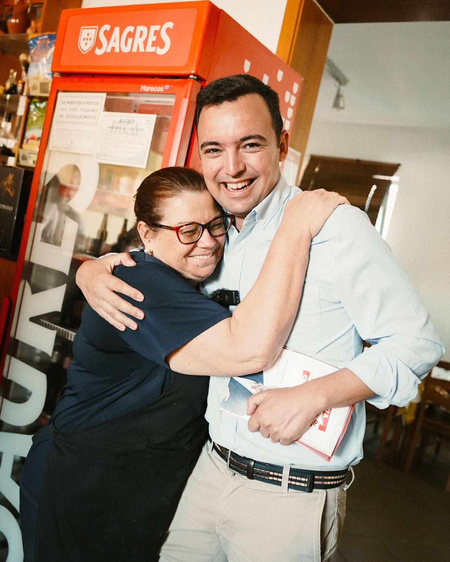 People embracing near a Sagres branded display in an indoor setting.