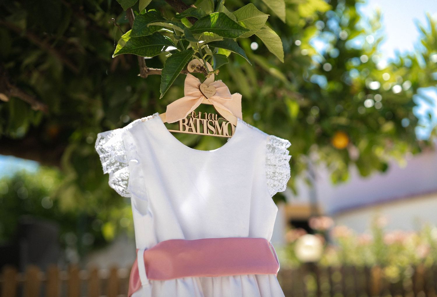 White flower girl dress on a wooden hanger labeled 'Flower Girl' hanging from a tree outdoors.
