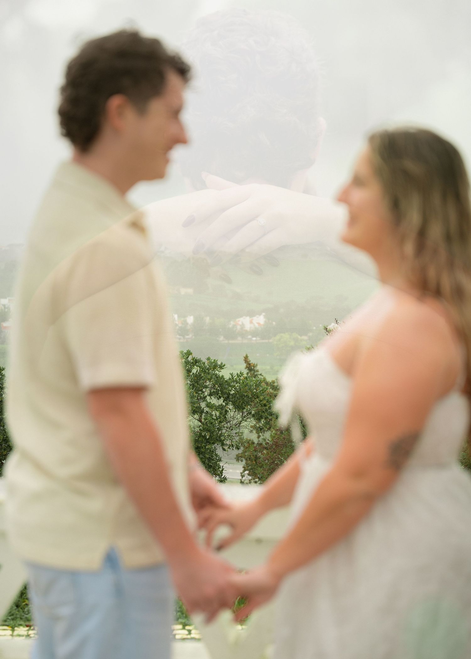 A couple holds hands facing each other in a dreamy double-exposure wedding photo with lush greenery backdrop.