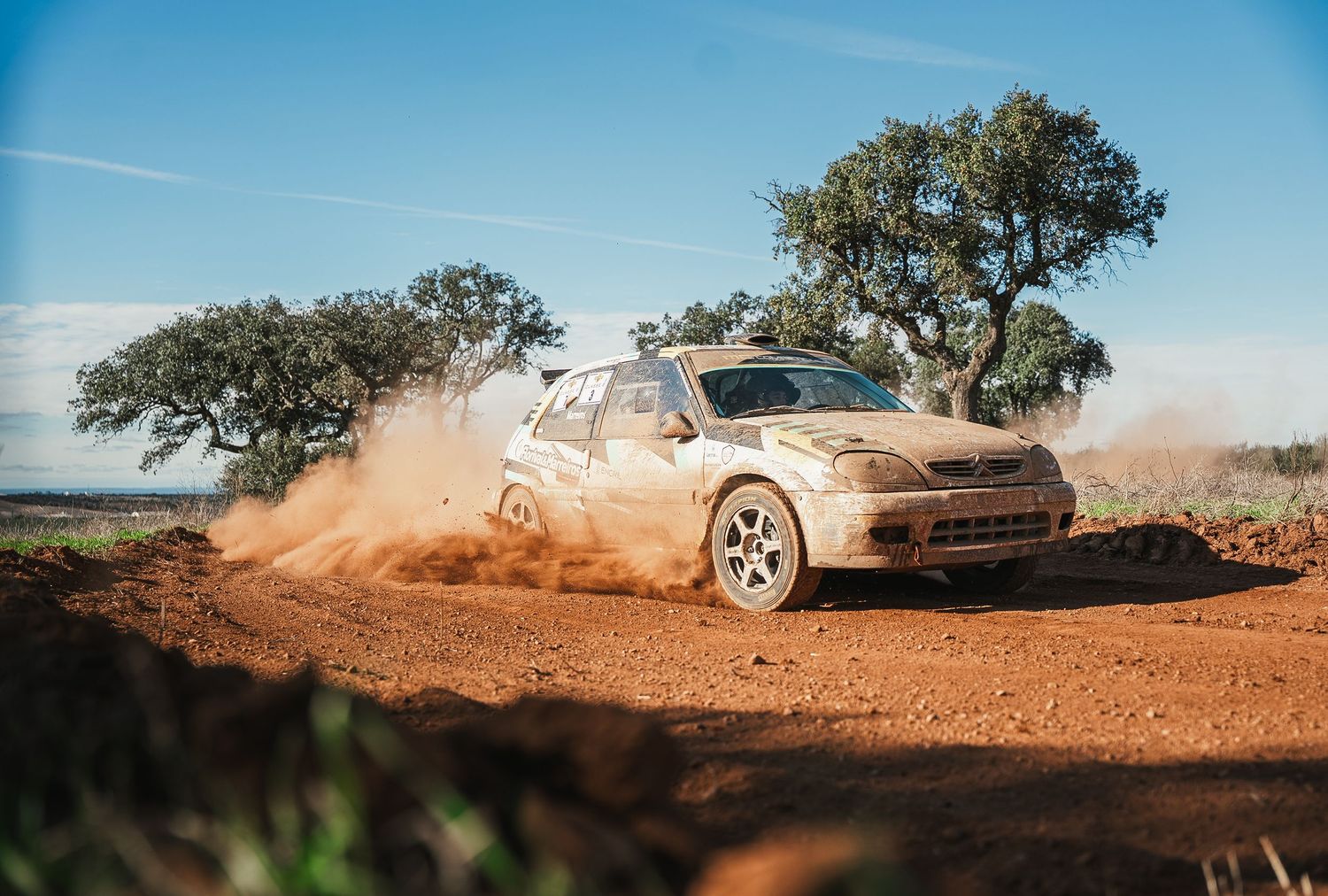 Rally car kicks up red dirt and dust while drifting around a corner during an off-road race.