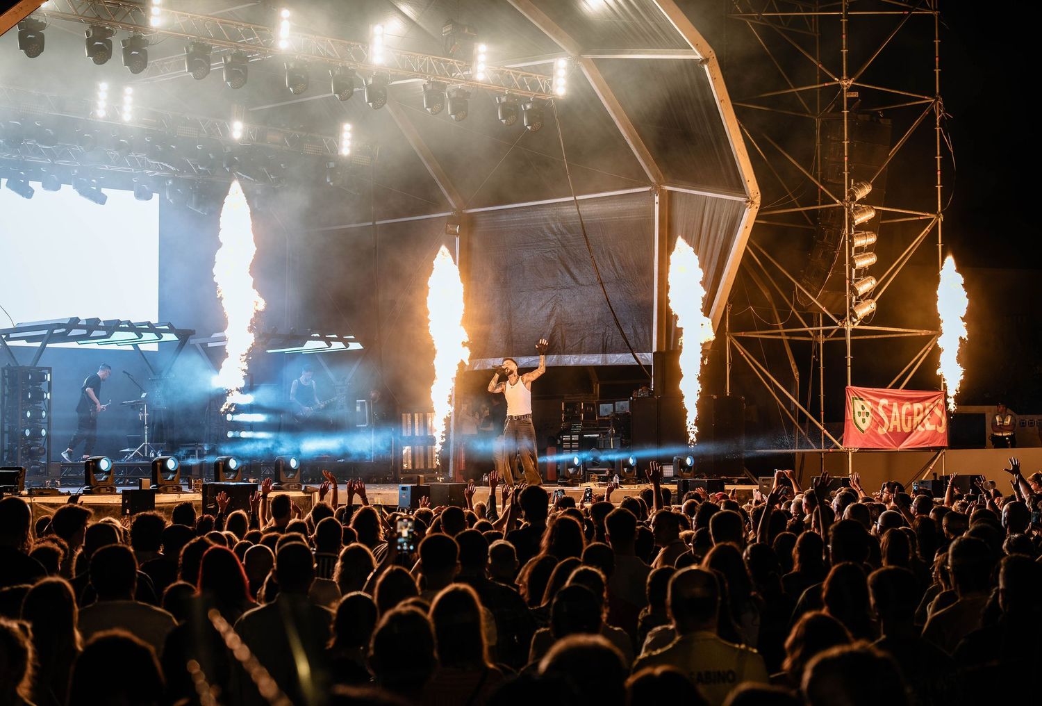 Large concert venue with pyrotechnic effects and crowd silhouettes beneath illuminated stage.