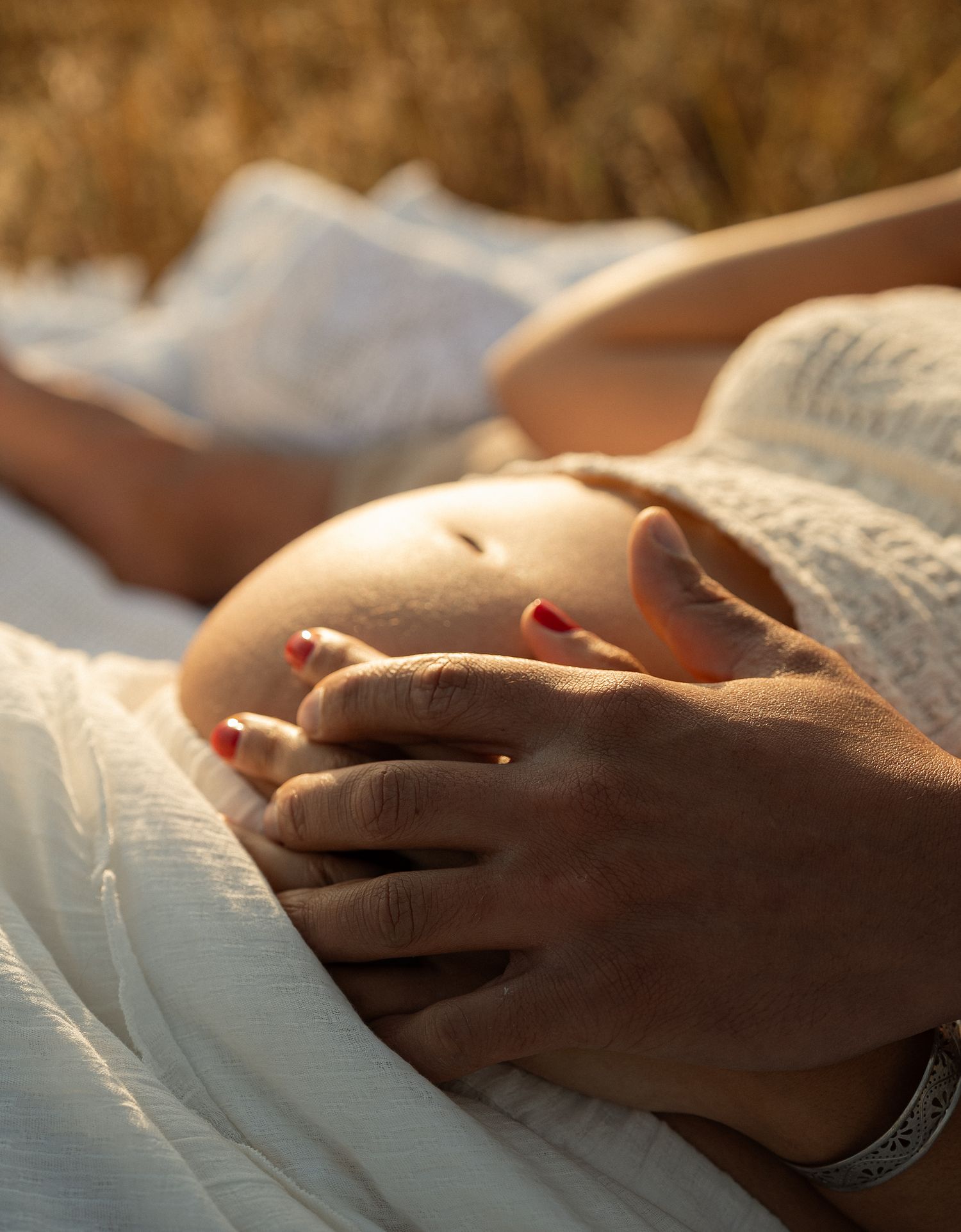 Pregnant woman in white dress lying in golden field, cradling baby bump with red-painted nails in warm sunlight.