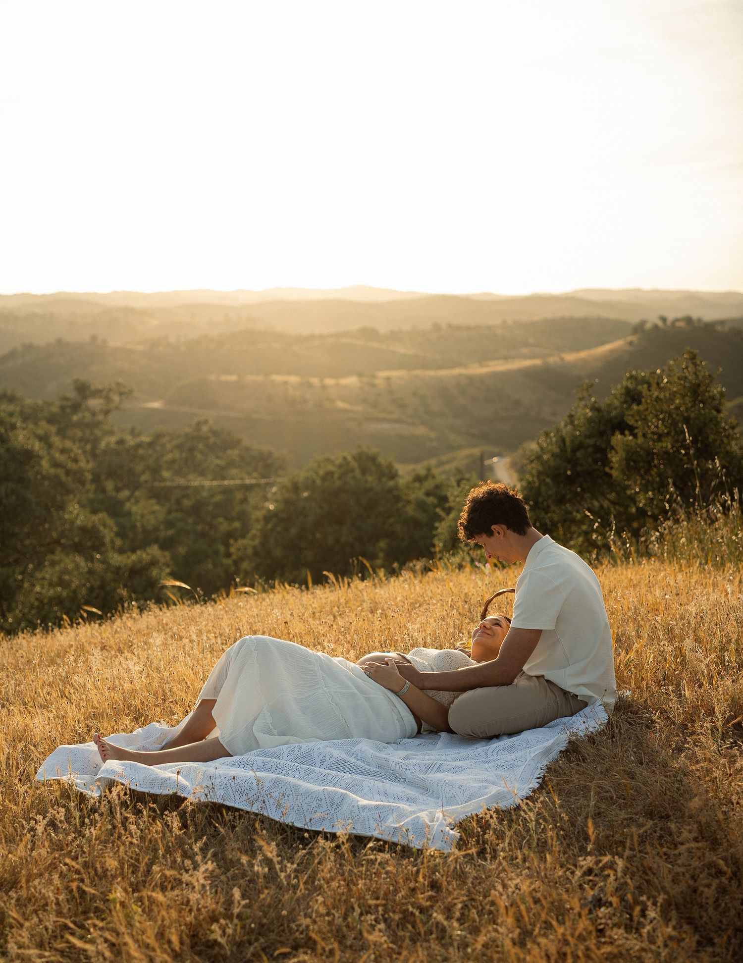 Couple relaxing on a blanket in golden fields at sunset with rolling hills in the background.