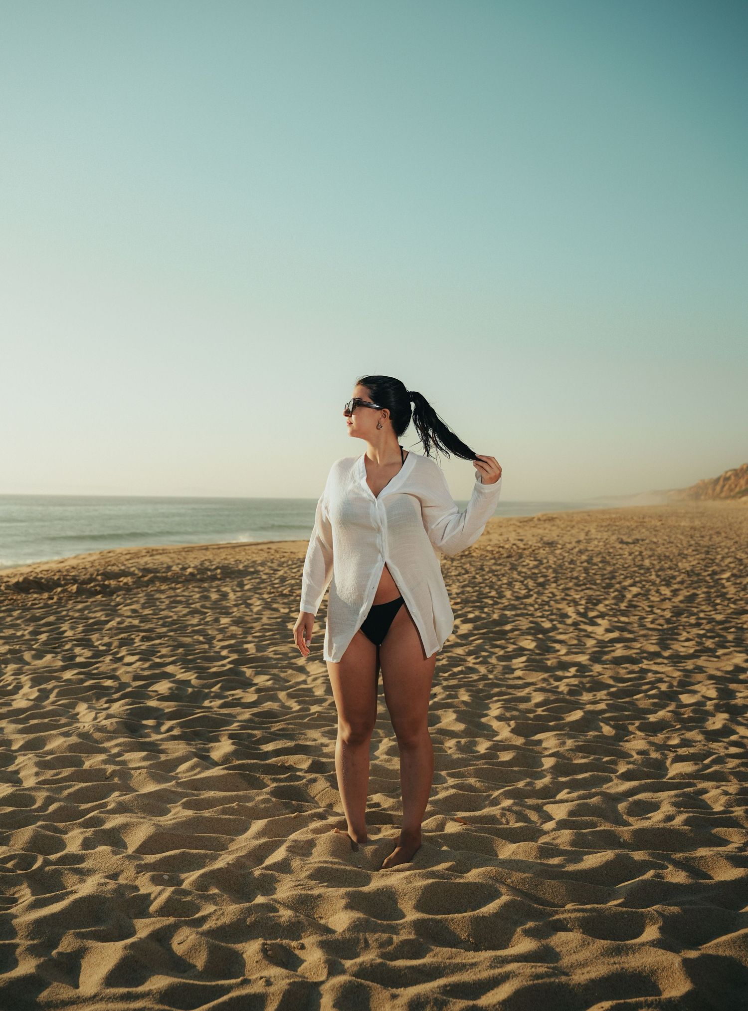 Woman in oversized white shirt poses on beach at golden hour.
