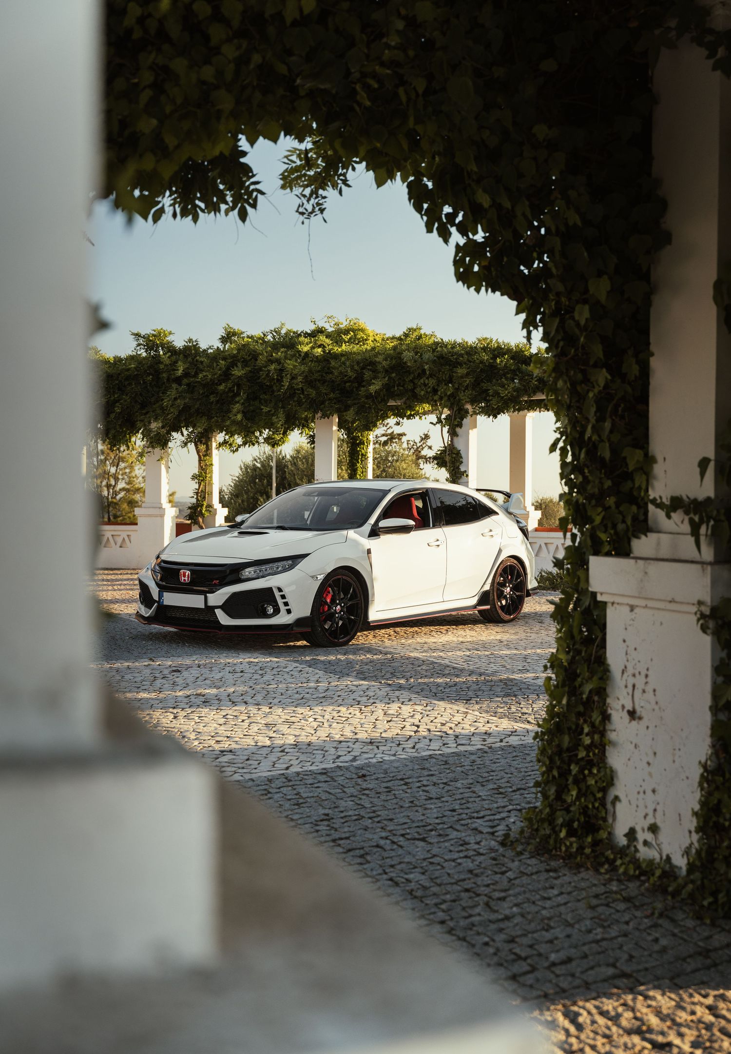 White sports car parked on cobblestone driveway with trees and architecture in background.