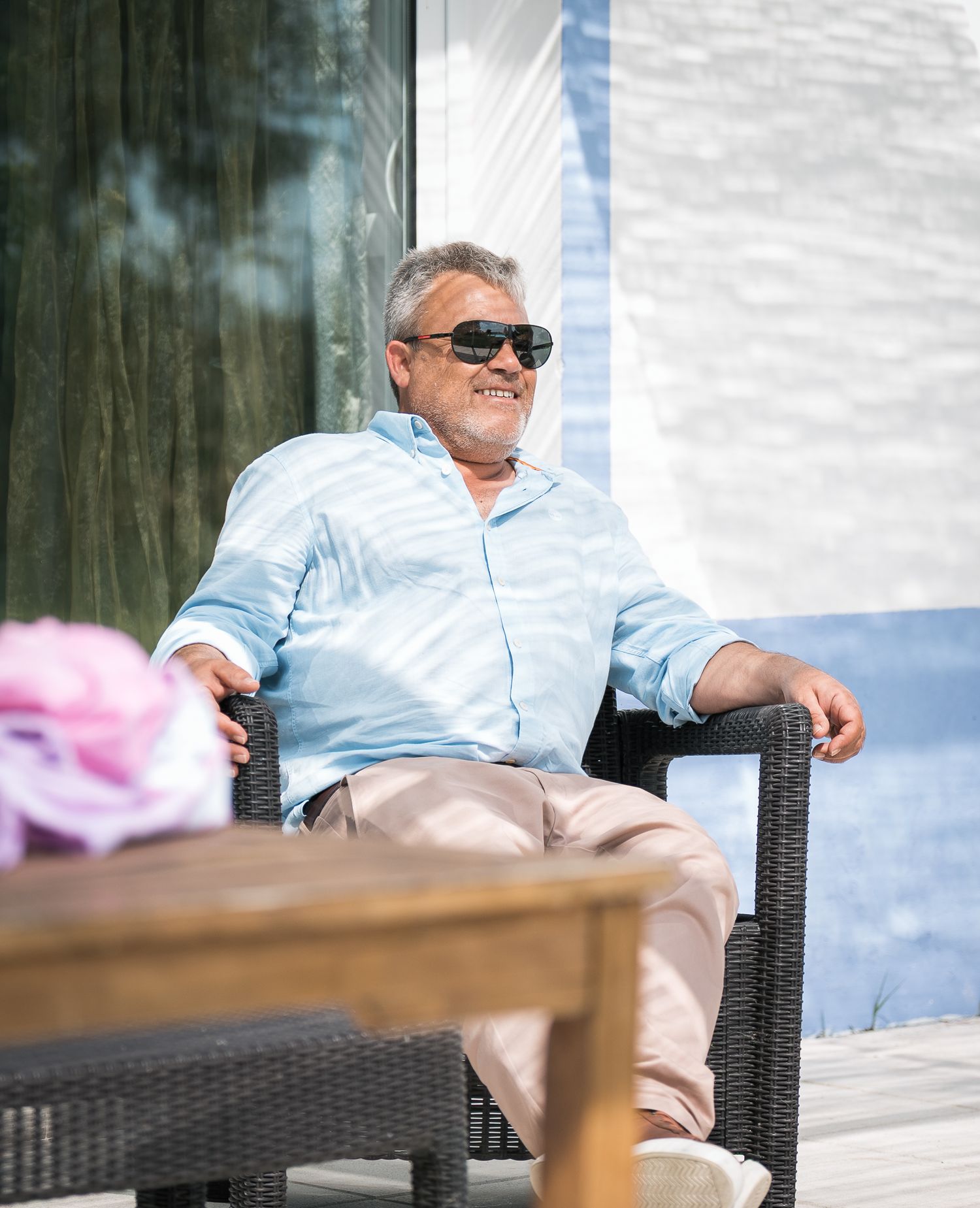 Person relaxing in wicker chair on sunny patio wearing blue shirt and sunglasses.