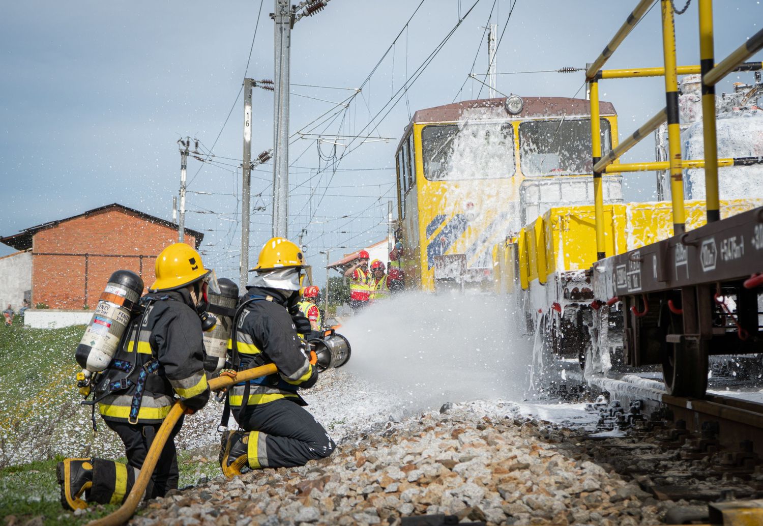 Firefighters in yellow protective gear working with equipment near train tracks.