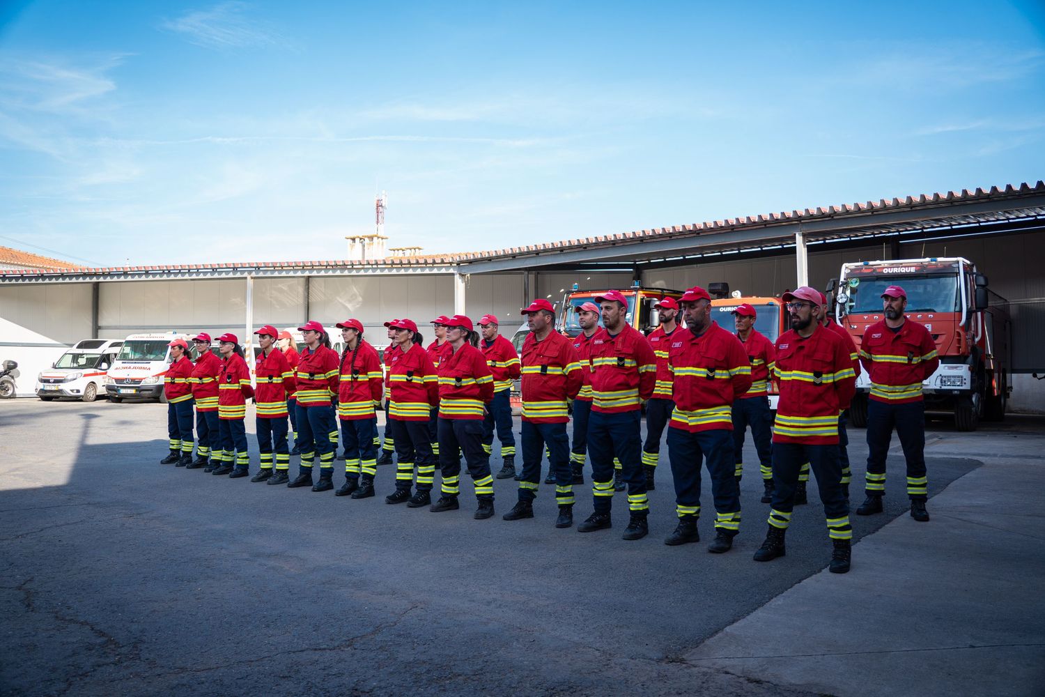Long line of firefighters in formation standing at attention on a sunny day outside their station.