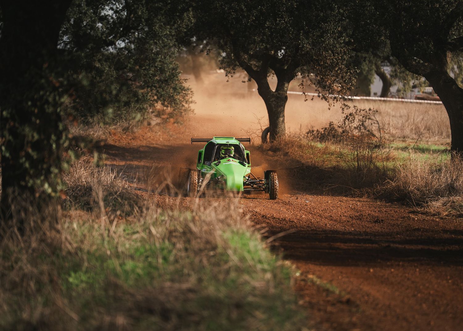 Off-road racing buggy kicking up dirt on a dusty trail between trees at dusk.