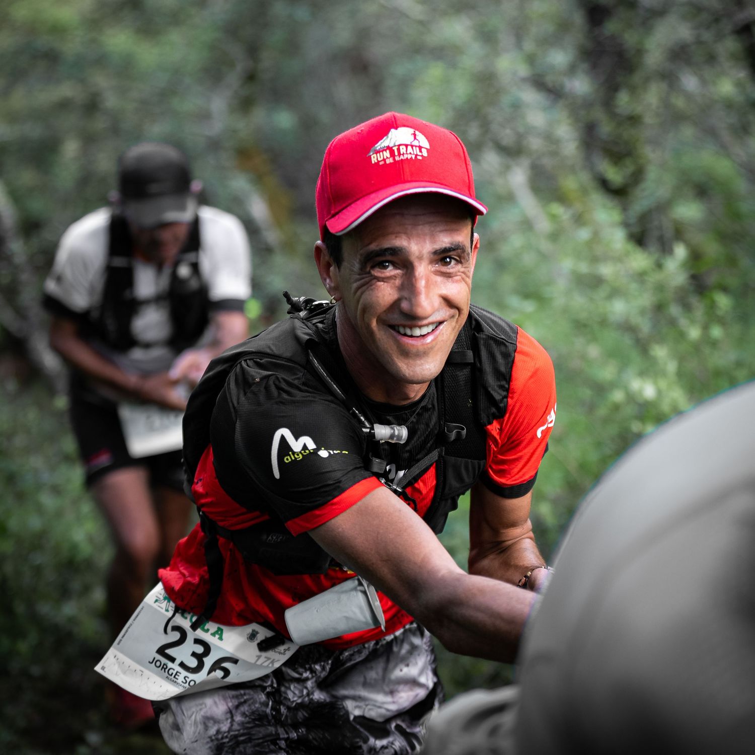 Trail runners wearing race bibs climb uphill through wooded terrain during a competition.