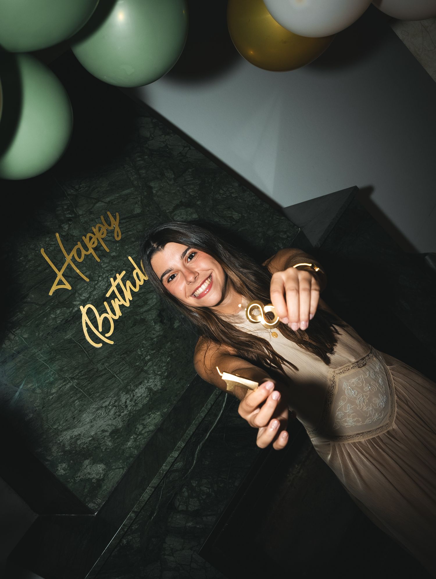 Woman in gray dress poses playfully against dark background with balloons and golden text overlay.