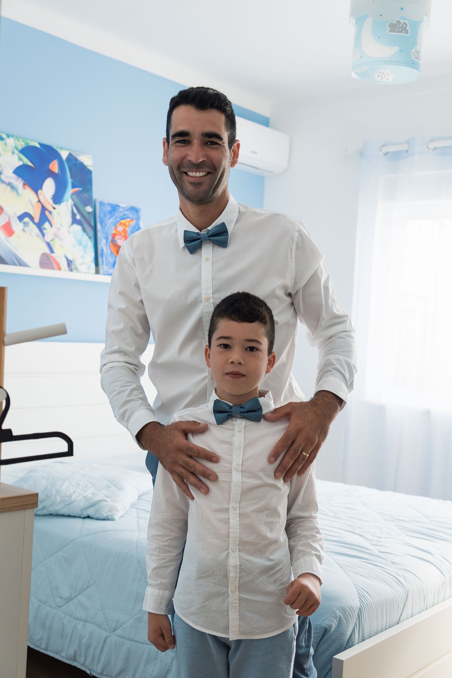 Two people in matching white shirts and blue bow ties posing together in a bedroom with Sonic artwork.