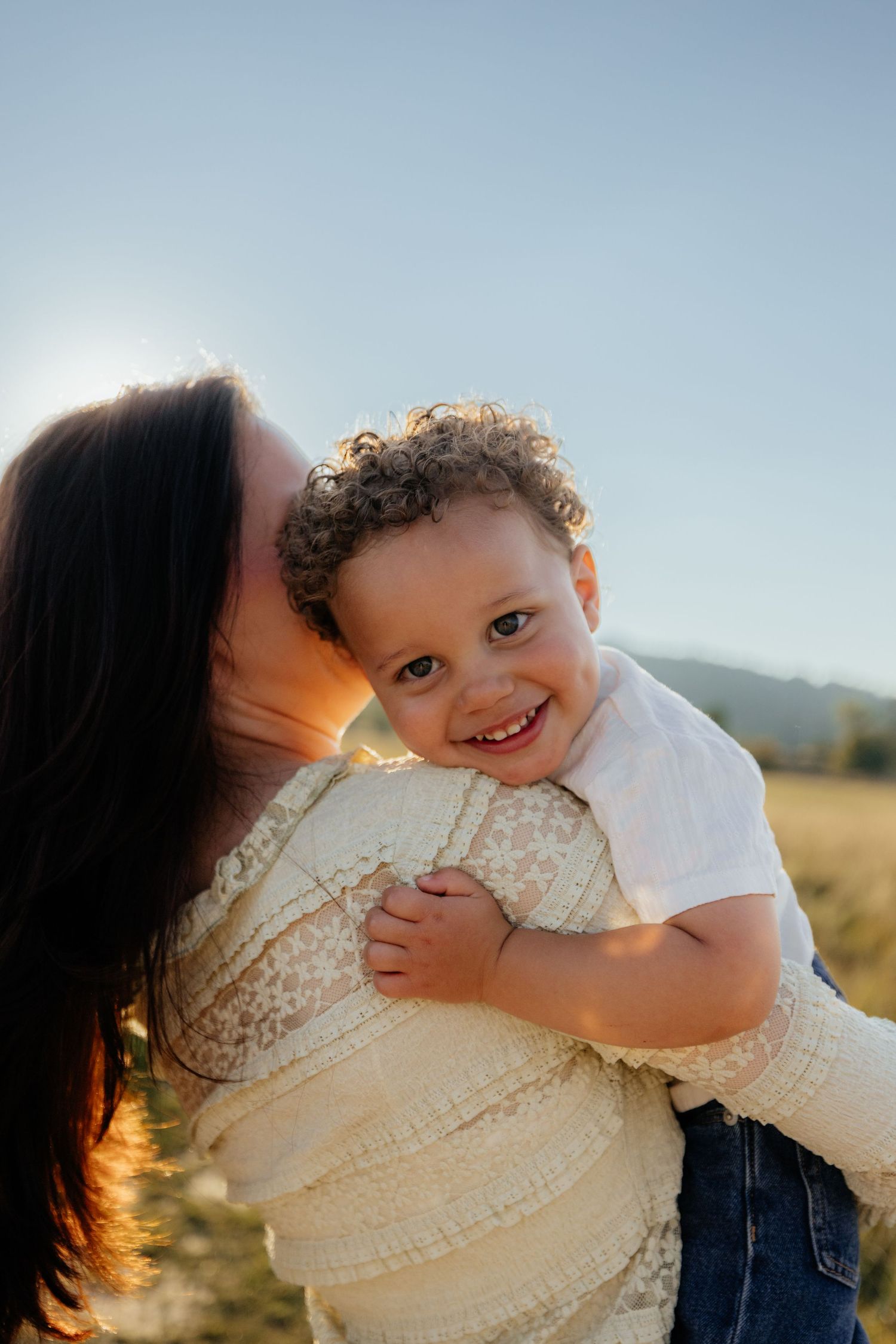 A mother kisses her smiling toddler with curly hair outdoors in golden sunlight.