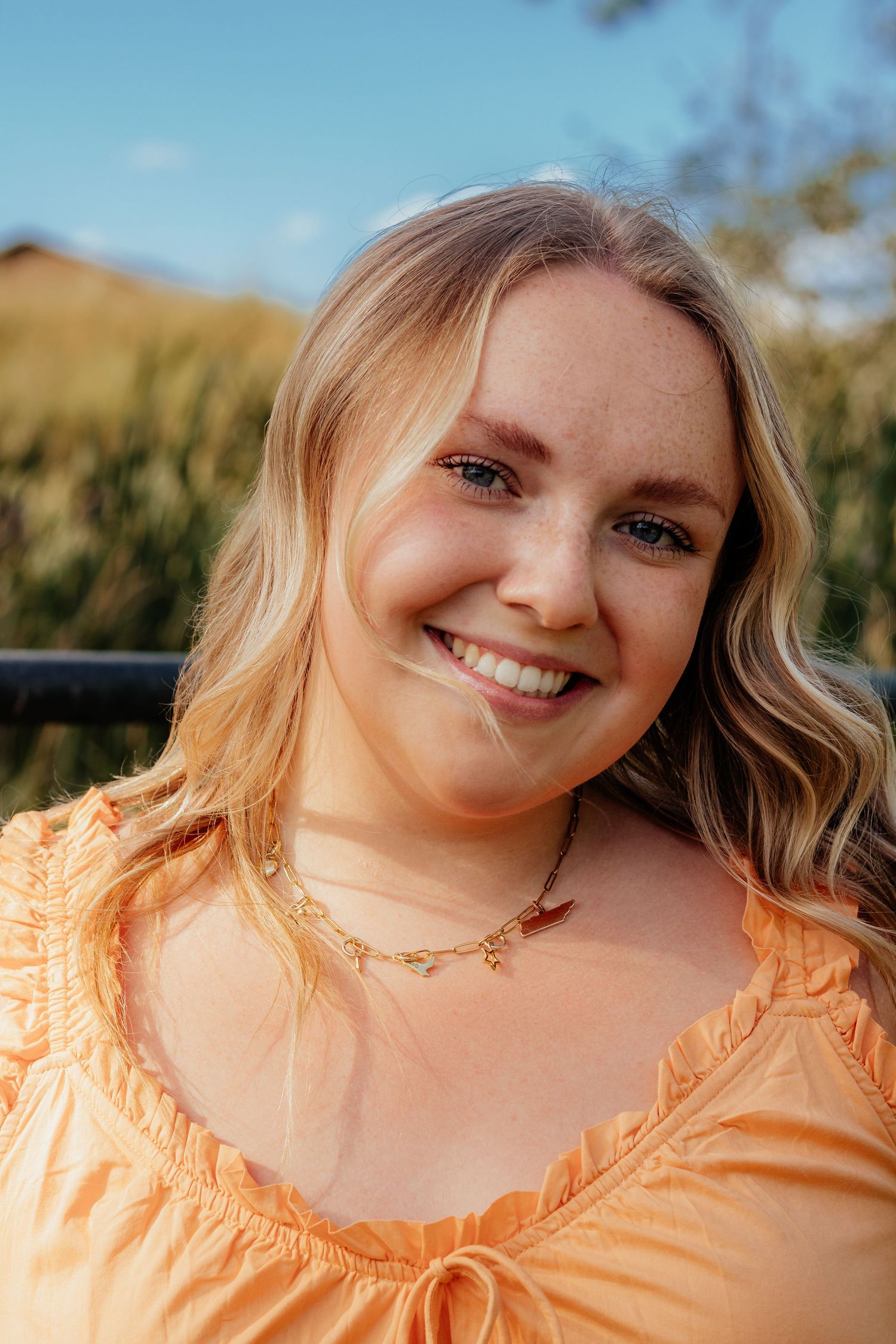 A person wearing a peach-colored top smiles warmly at the camera in an outdoor setting with golden sunlight.