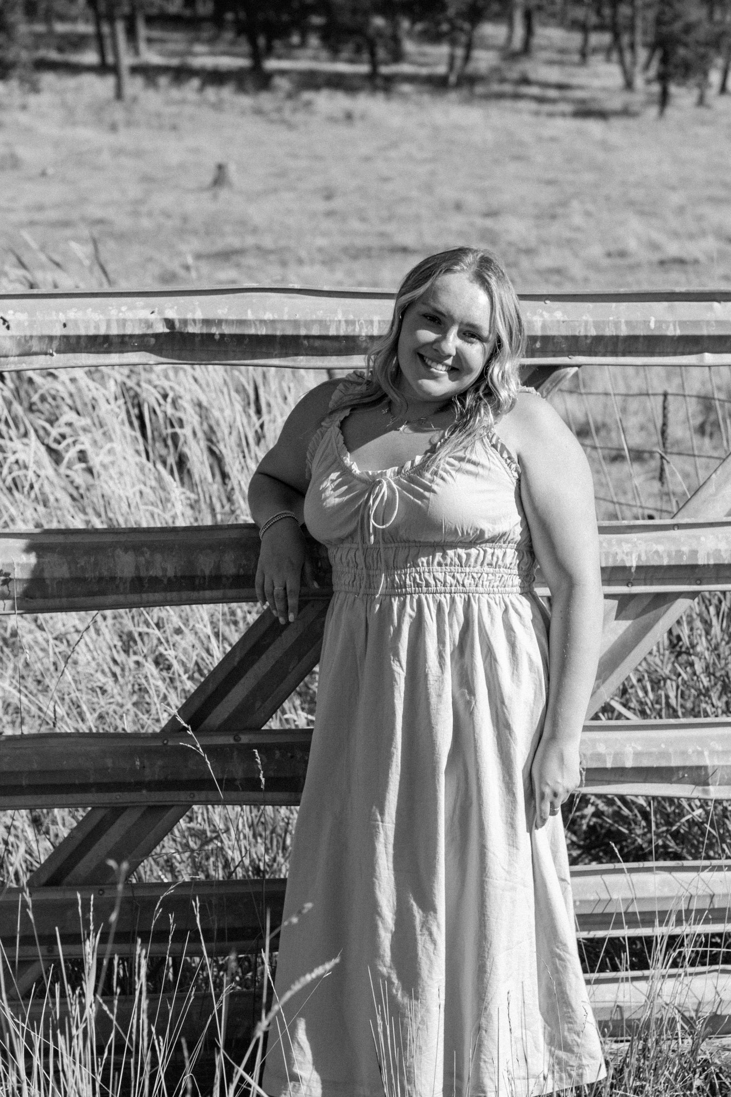 Person in a white sundress poses near a rustic wooden fence in black and white.