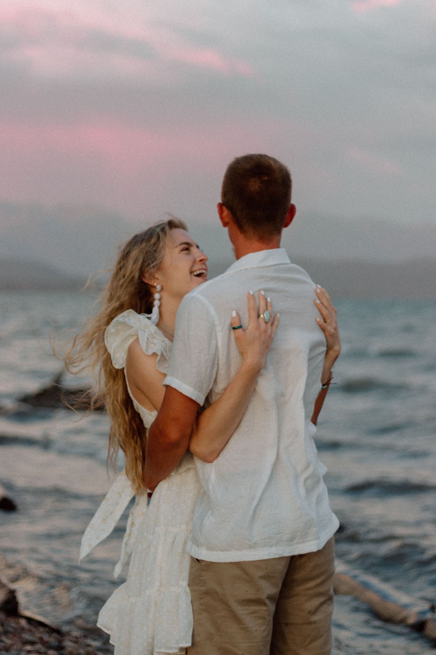 A romantic couple laughs and embraces on a beach during sunset with waves crashing behind them.