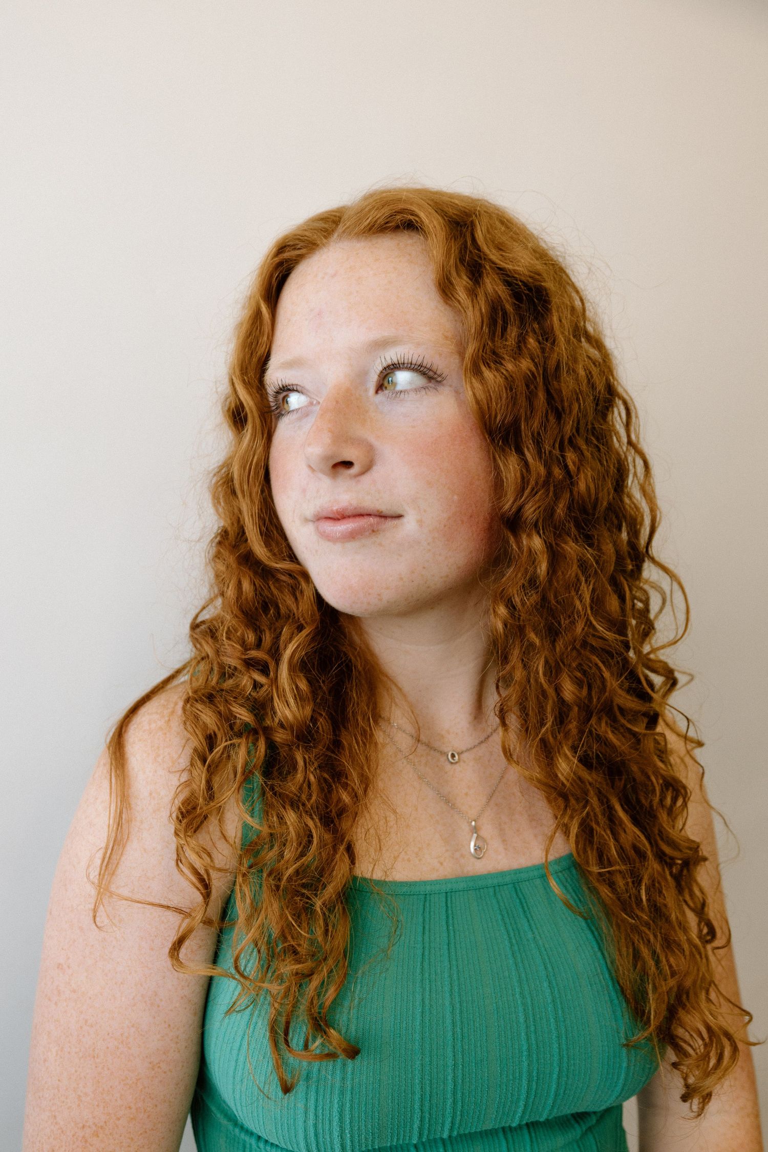 Person with curly red hair poses playfully in green top against white wall.