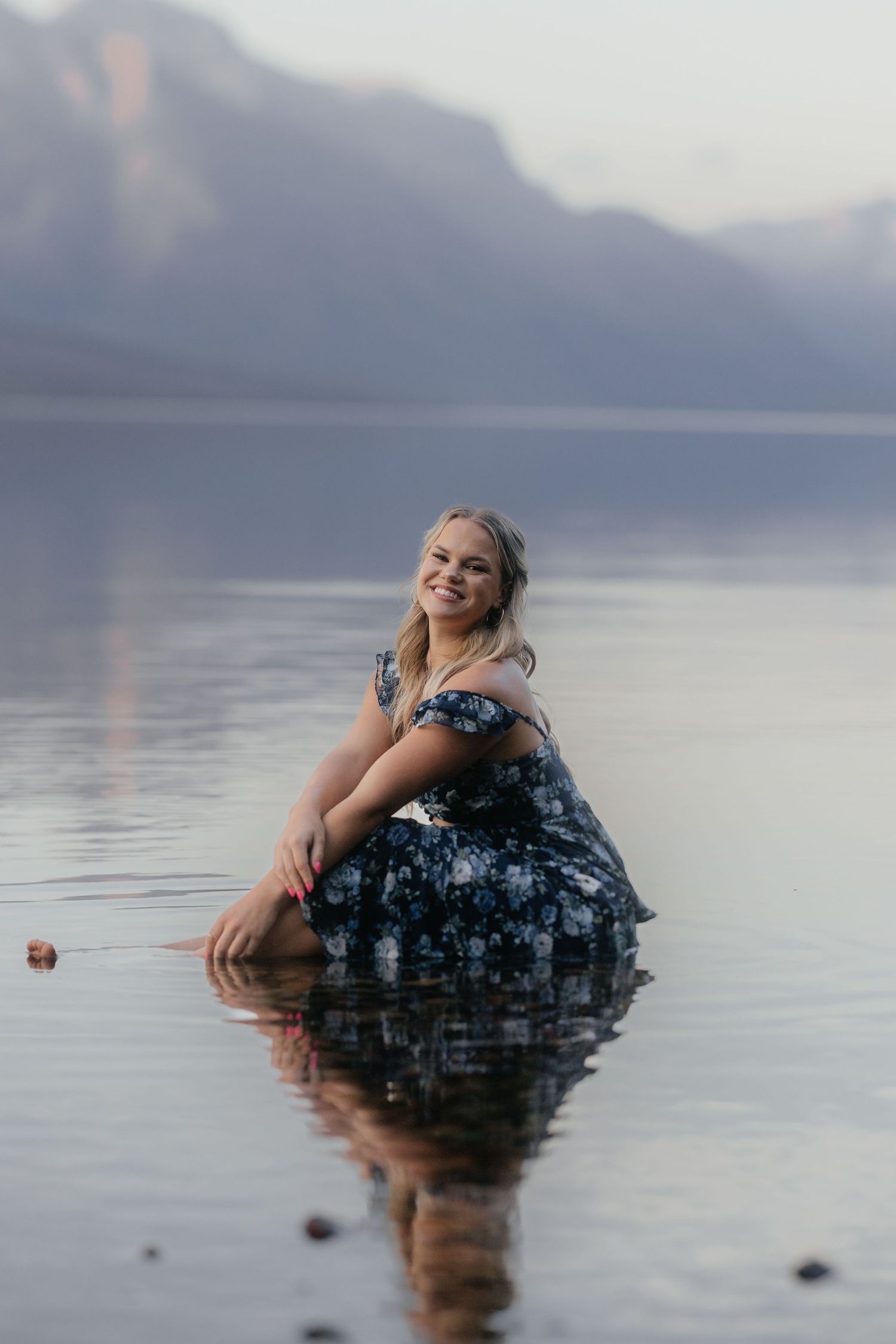 A person wearing a dark floral dress floats serenely in calm lake waters at dusk with mountains in the background.