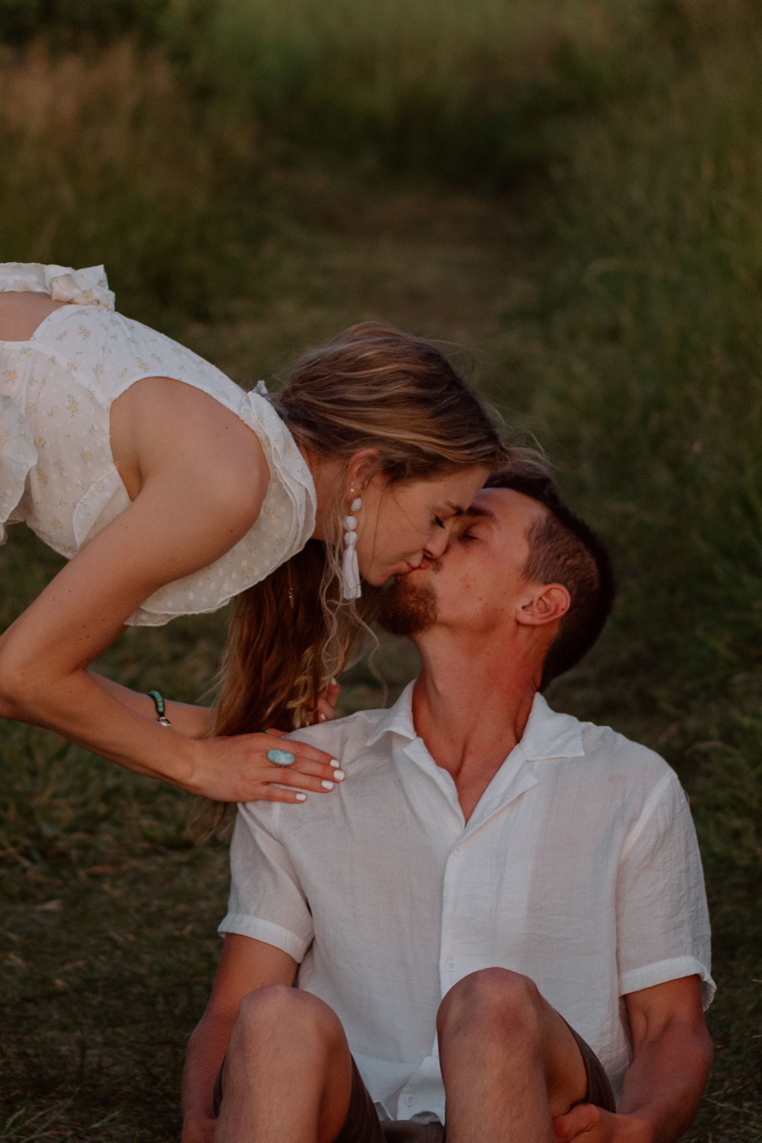 A romantic couple in white outfits share tender moments while sitting together outdoors at sunset.