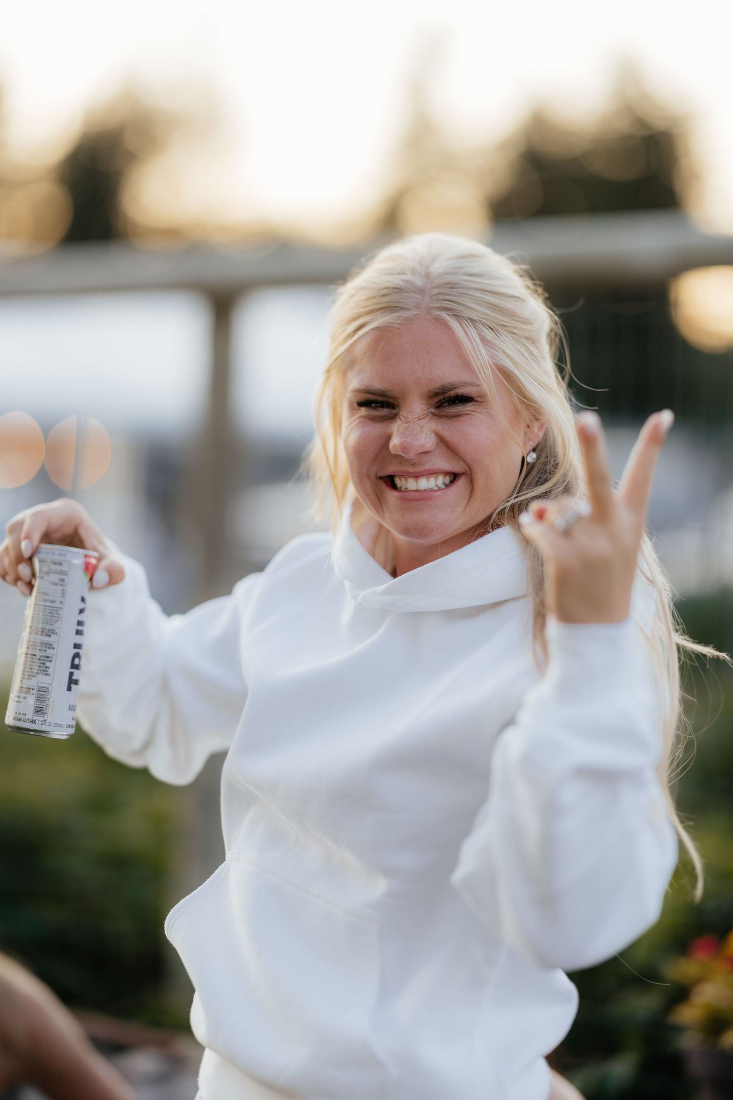 A person in a white sweater laughs joyfully while holding up their arms in a sequence of celebratory poses outdoors.