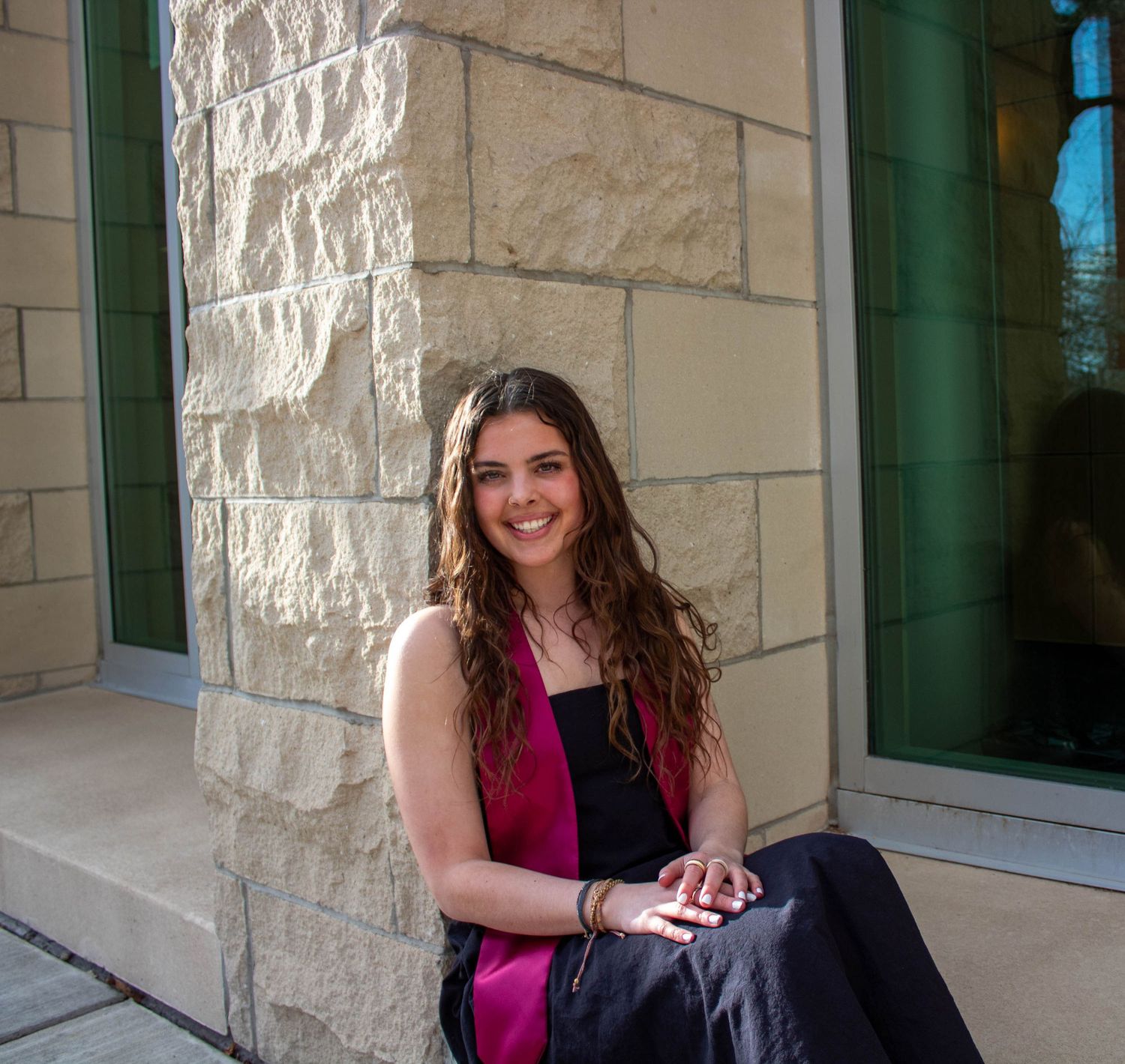 Woman in magenta dress relaxes on stone exterior steps near green-framed windows and light stone walls.