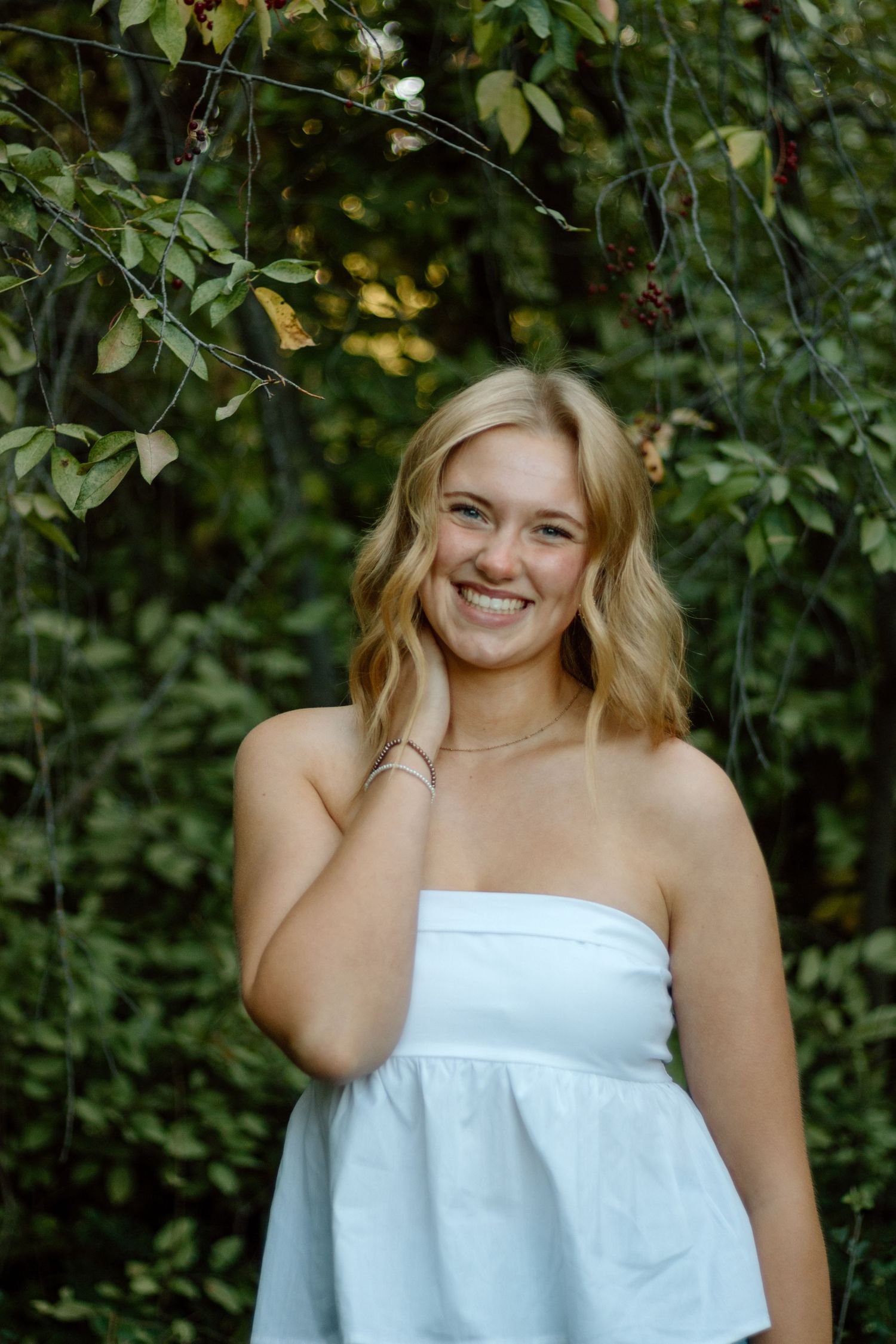 A person in a white strapless top poses outdoors against a leafy green background.