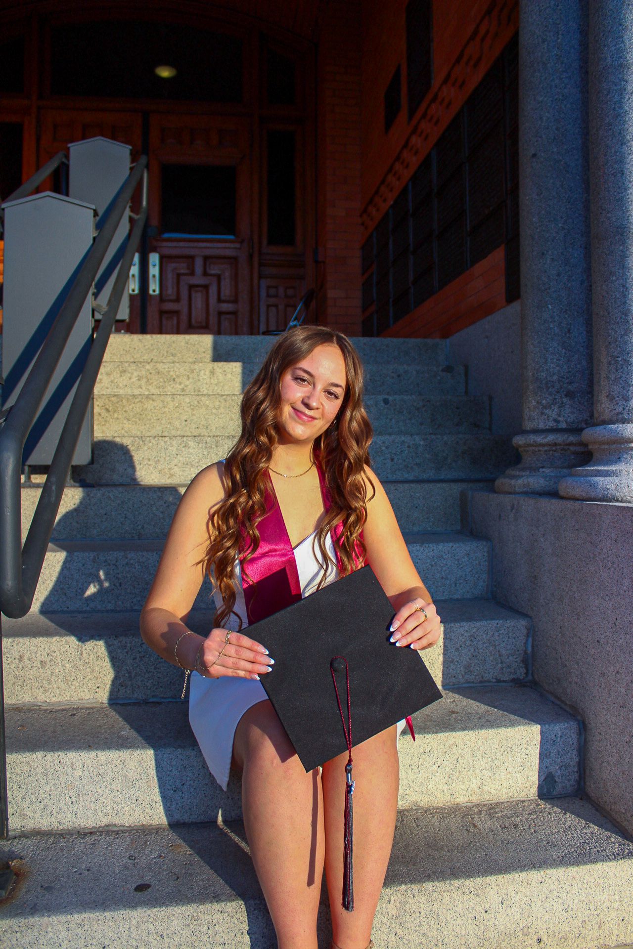 A graduate sitting on stone steps while smiling and holding a diploma during a sunset graduation photoshoot.