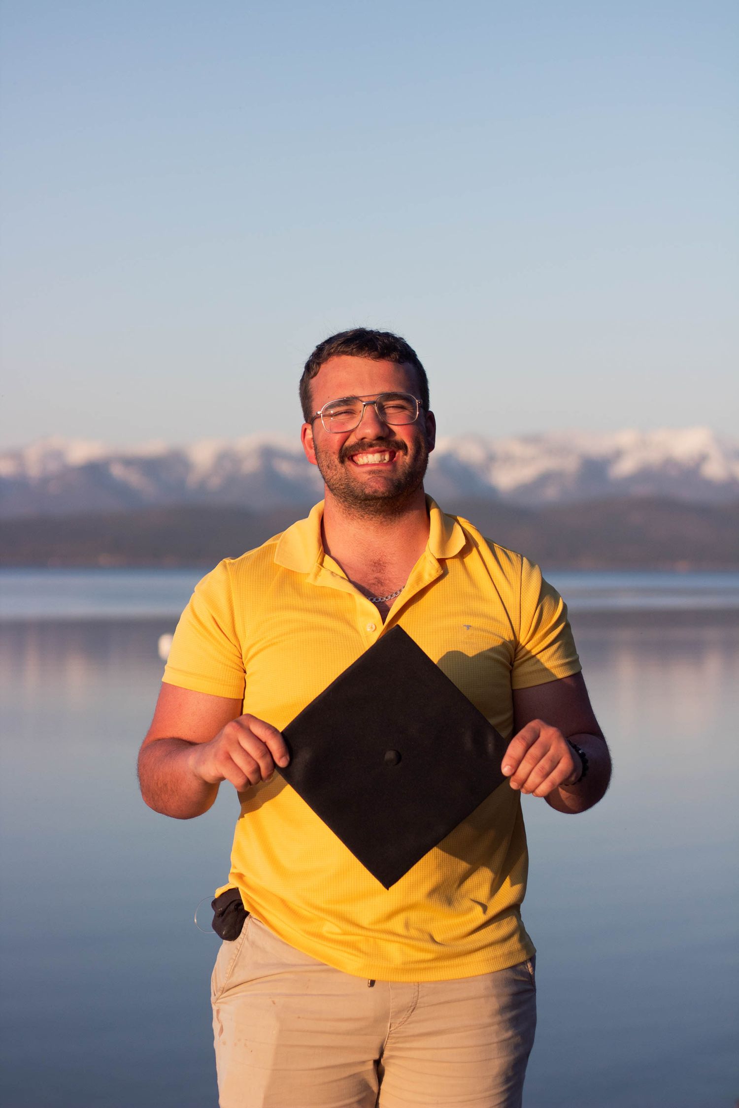 Someone in a yellow polo shirt holds a graduation cap by a tranquil lake with snow-capped mountains in the background.