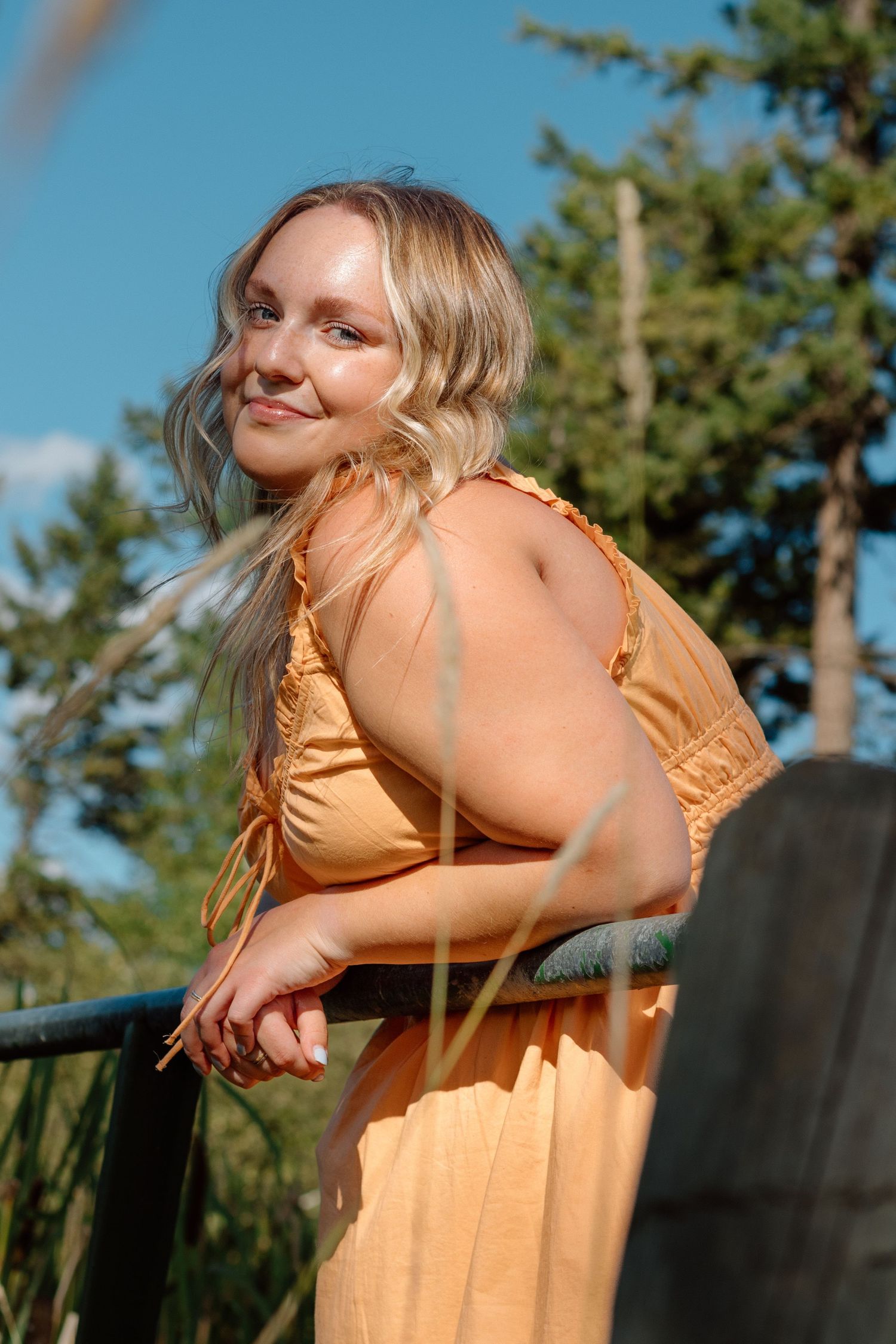 Someone wearing a flowing peach-colored dress leans against a railing in golden sunlight with evergreen trees behind.