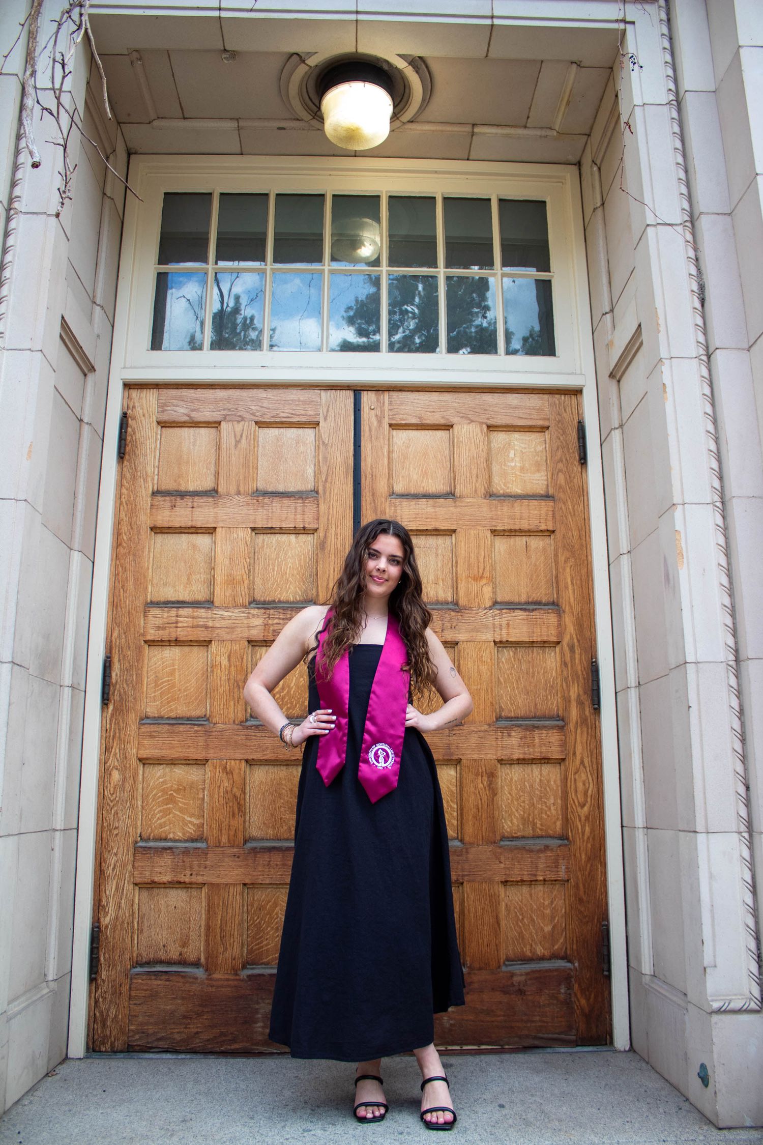 A person wearing a black maxi dress and pink sash poses in front of large wooden doors in an elegant entryway.